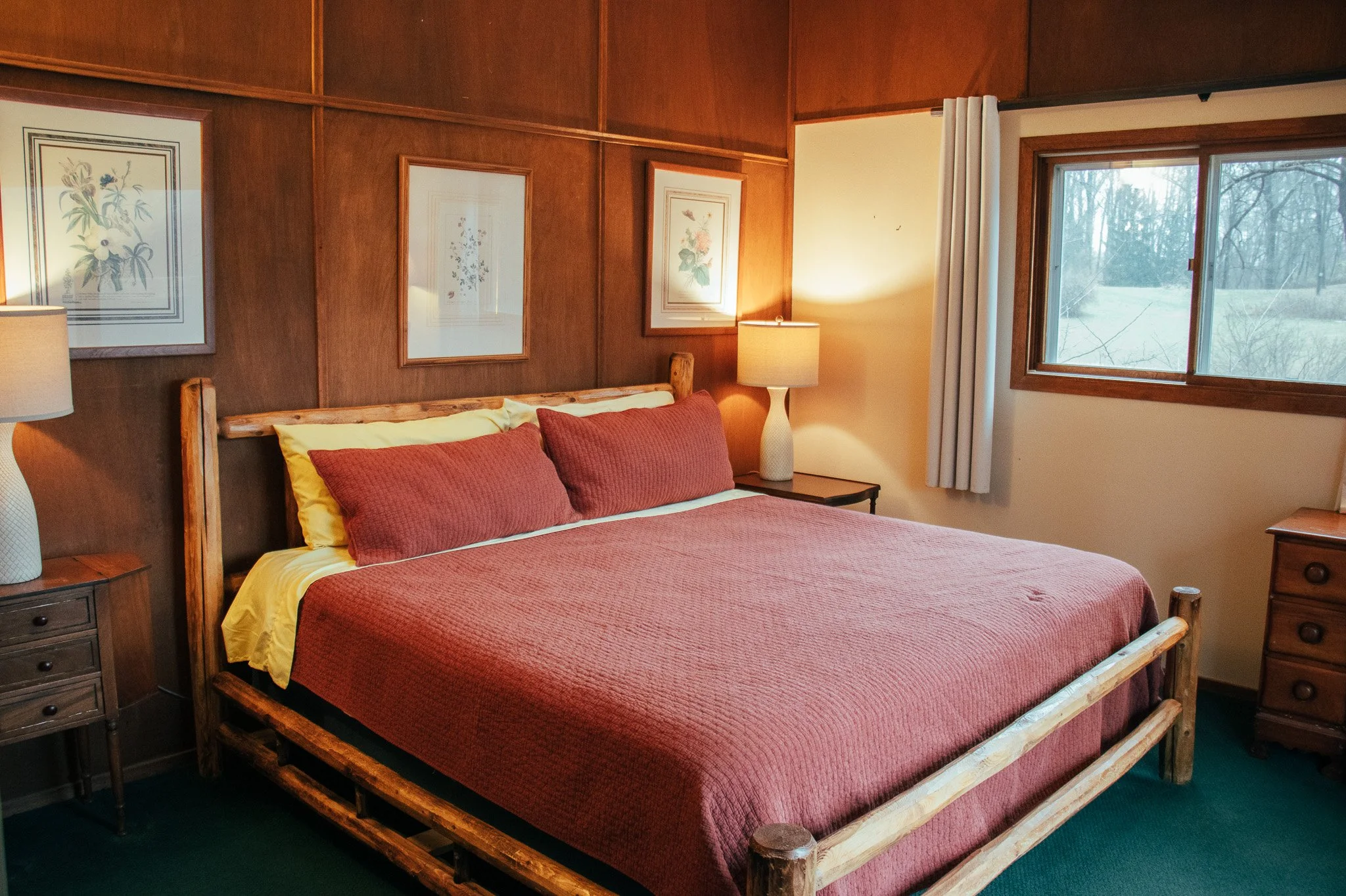 A cozy bedroom with wood-paneled walls, a large bed with rust-colored bedding and pillows, two bedside tables with lamps, framed botanical art prints on the wall, a window with white curtains showing an outdoor scene with trees.