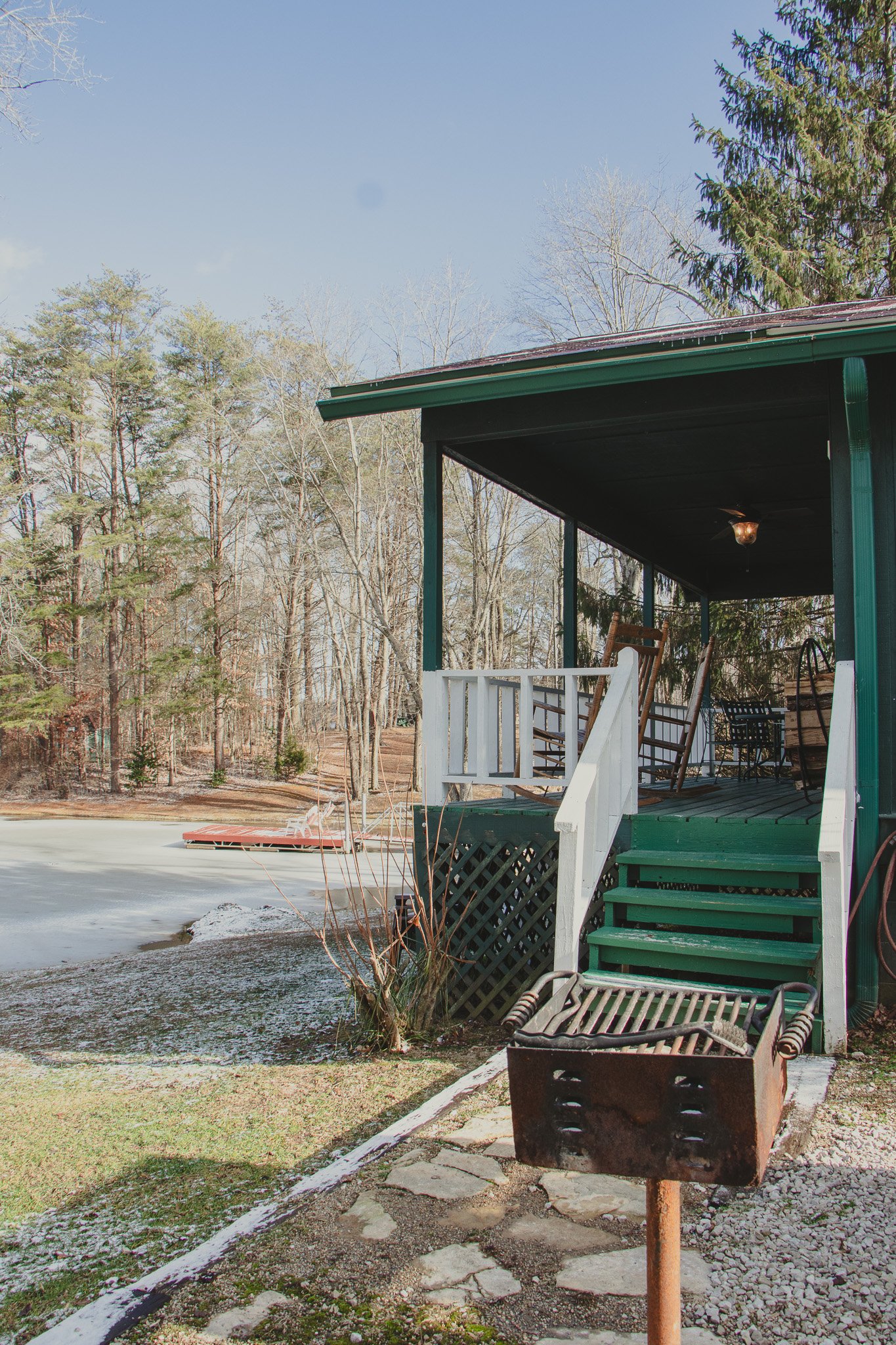 A wooden house with a porch overlooking a frozen pond, with trees in the background during winter.