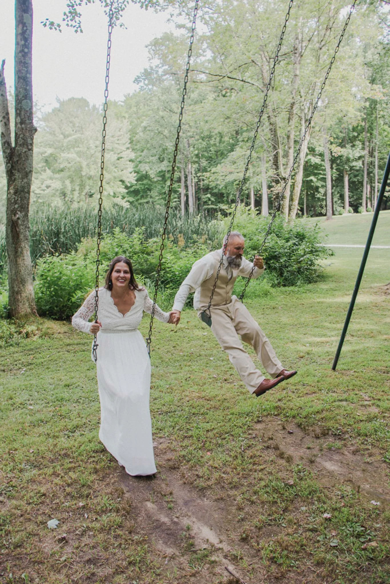 A woman in a white wedding dress and a man in beige clothing are on a park swing, holding hands, surrounded by trees and greenery, smiling and appearing joyful.