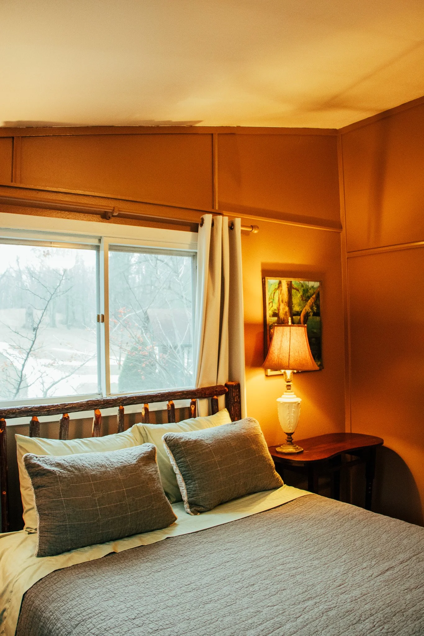 A cozy bedroom with a wooden bed frame, two pillows, beige bedding, a bedside table, a lamp, a framed picture, beige walls, and a window with curtains showing frosty trees outside.