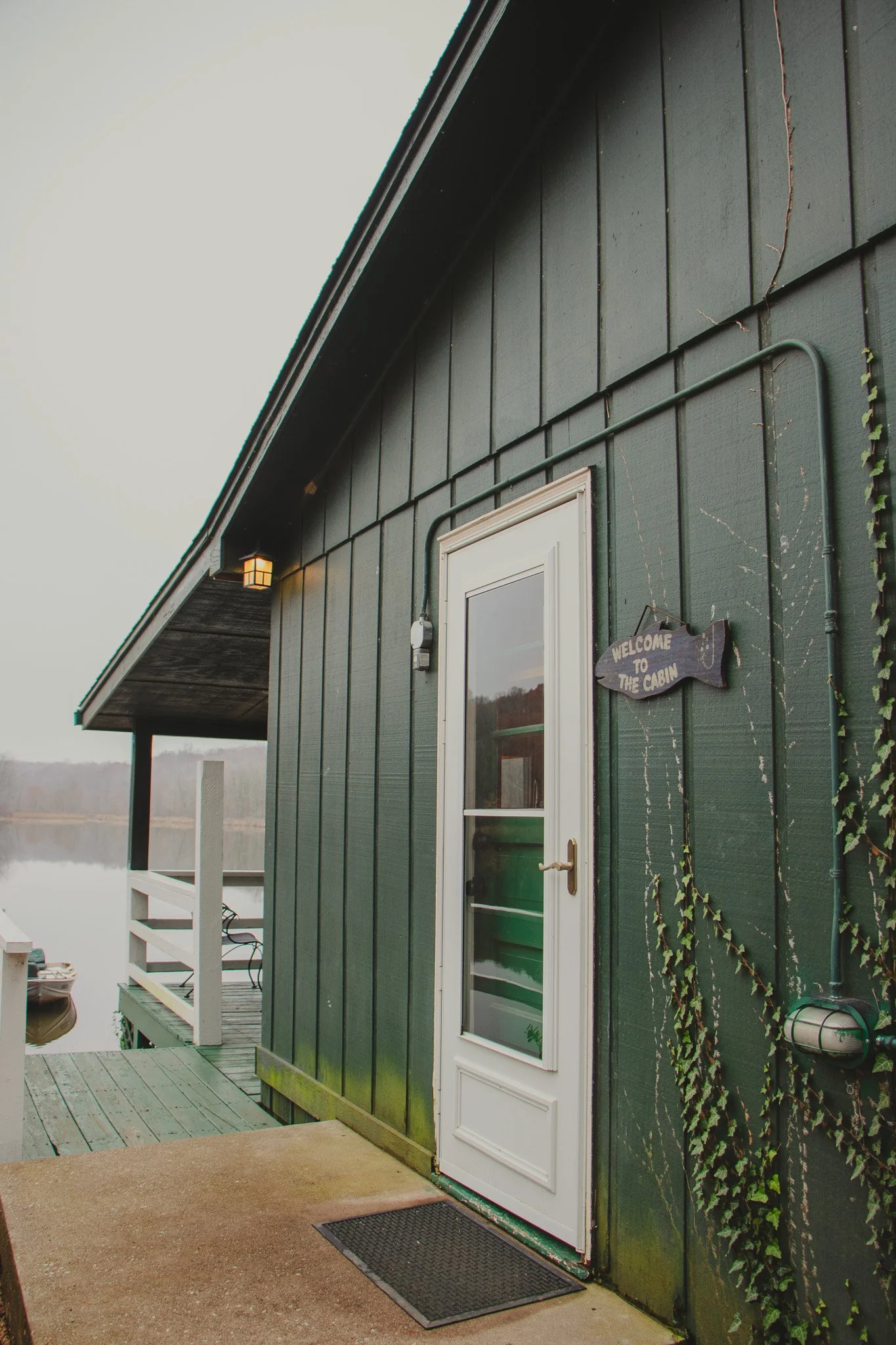 Green cabin with a white door, a wooden "Welcome to the Cabin" sign shaped like a fish, and an outdoor light fixture, situated near a lake.