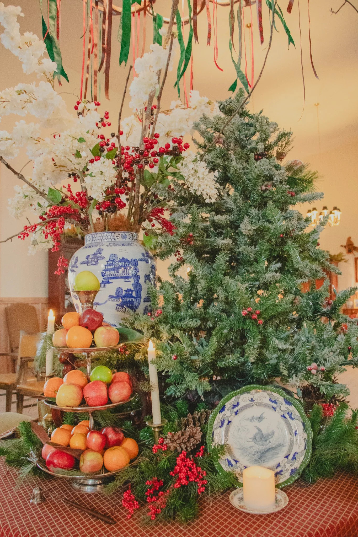 A decorated holiday table with a Christmas tree, fruit display, floral arrangement, candles, and decorative plates.
