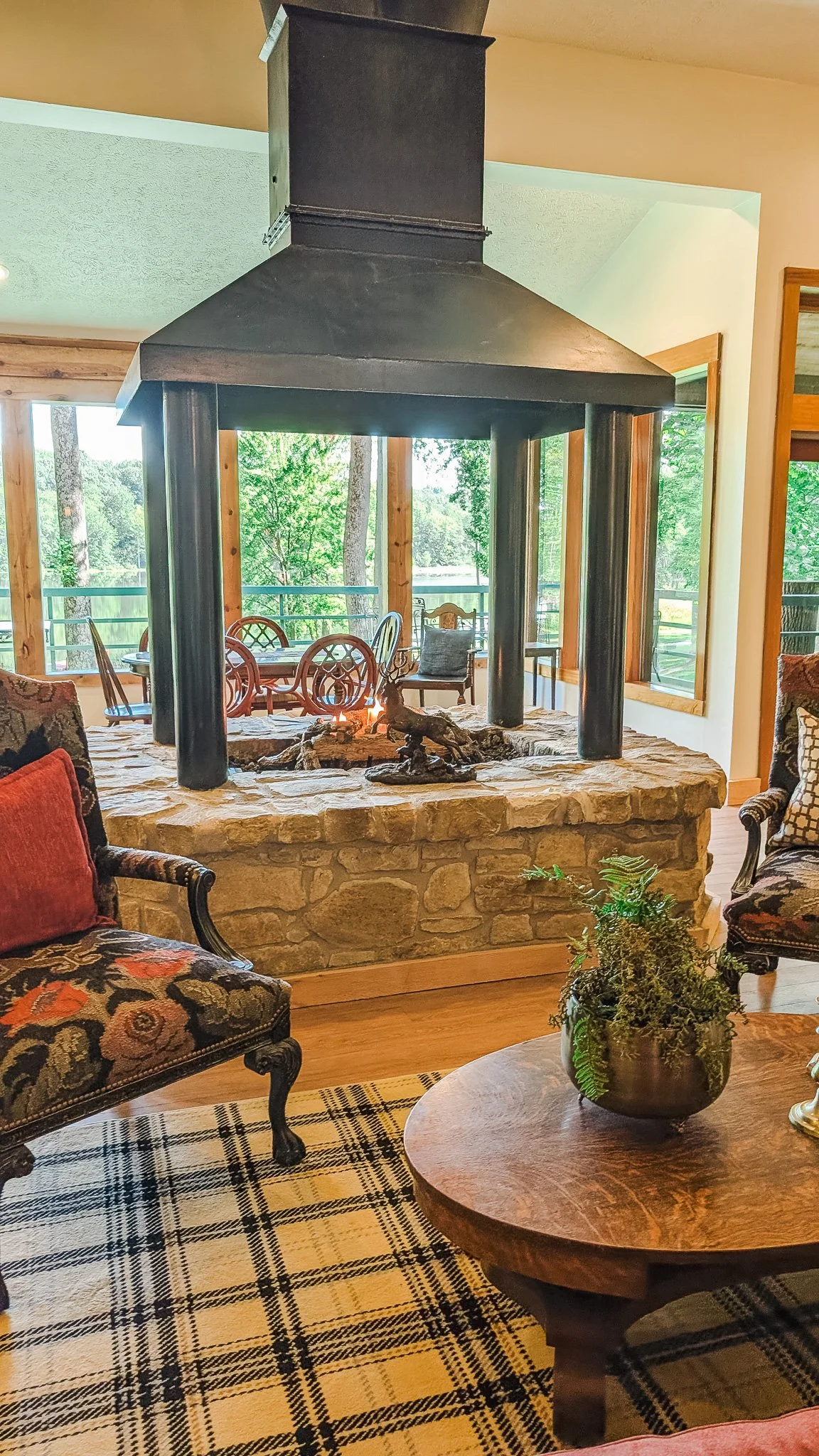 A cozy living room with a stone fireplace and a large black metal chimney hood, surrounded by wooden furniture, large windows revealing a green outdoor area, and decorated with a patterned rug and plants.