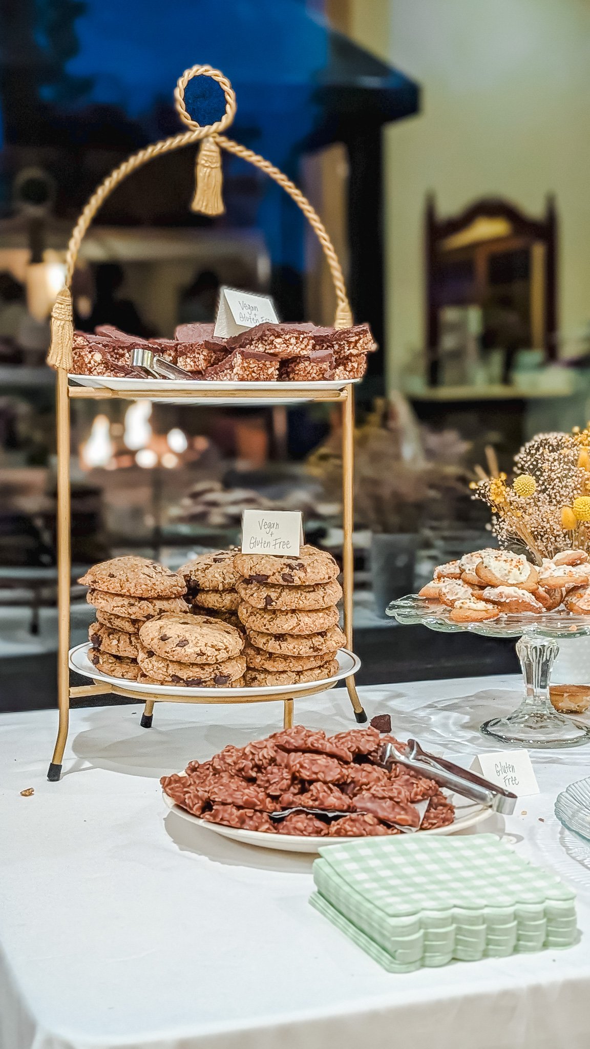 Assorted cookies on display at a dessert table, including vegan, gluten-free options, with a tiered stand and decorative flower arrangement.