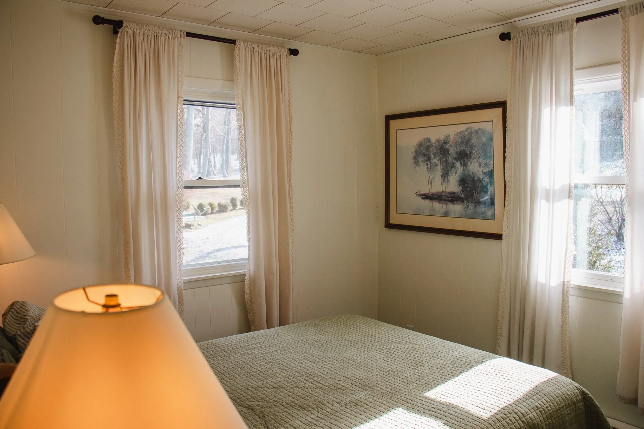 A bedroom with a bed covered in a beige quilt, two windows with cream curtains, a framed painting of trees on the wall, and a lamp with a beige shade in the foreground.