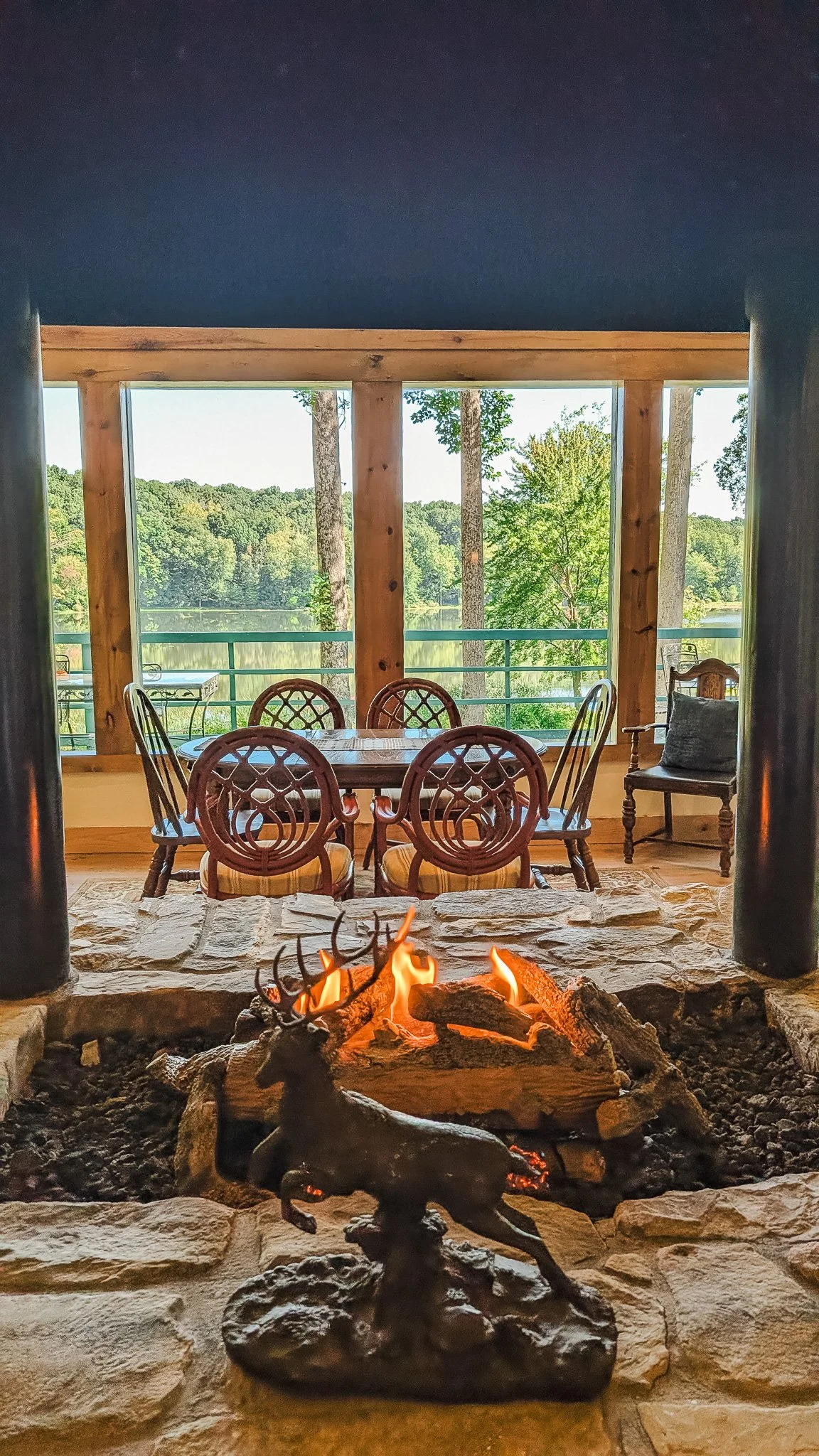 View from a fire pit with a deer sculpture, looking out onto a screened porch with a dining table and chairs, overlooking a lake and green trees.