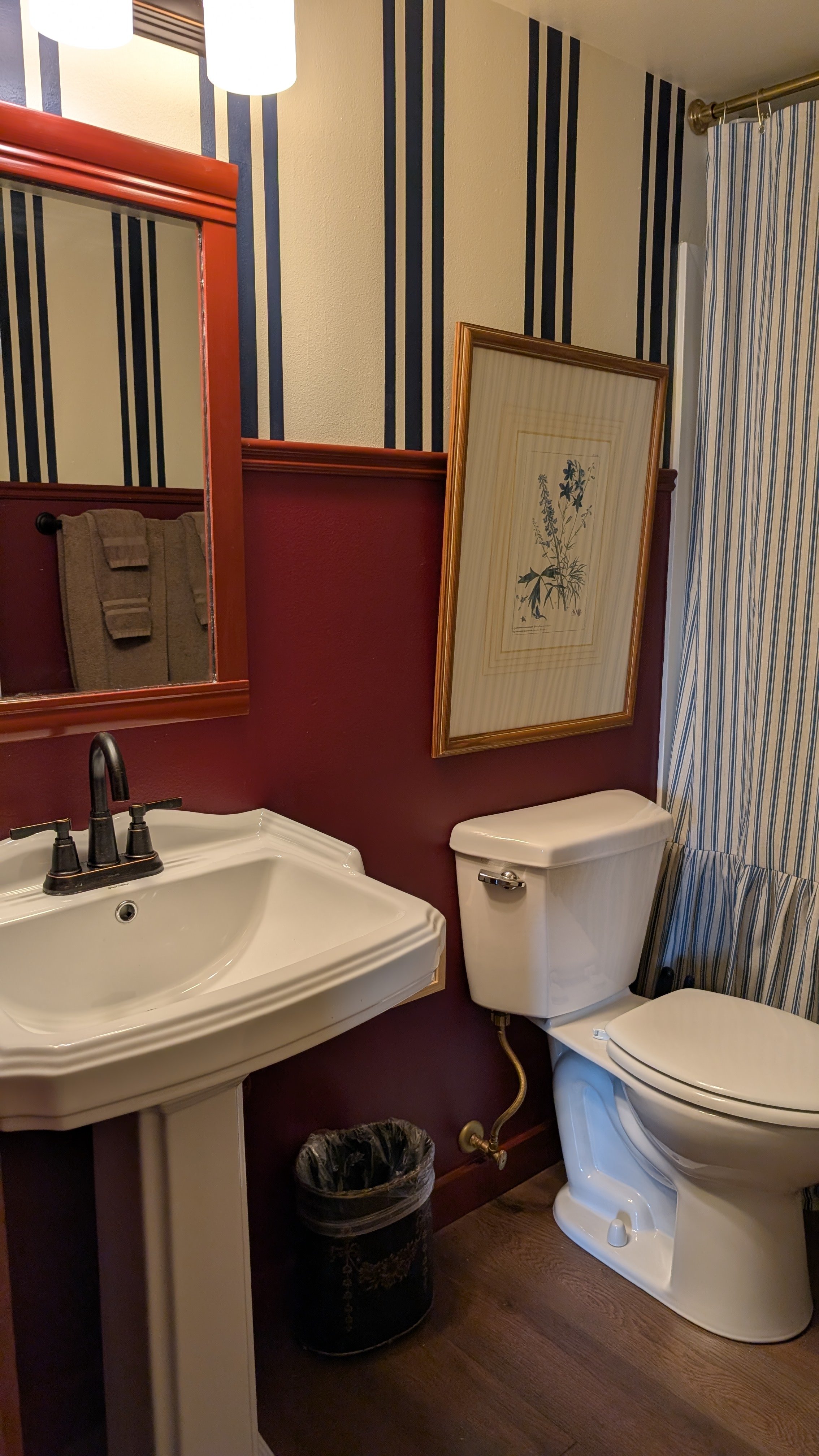 Upstairs bathroom with navy striped walls, red wainscotting, blue tick shower curtain and floral art print.