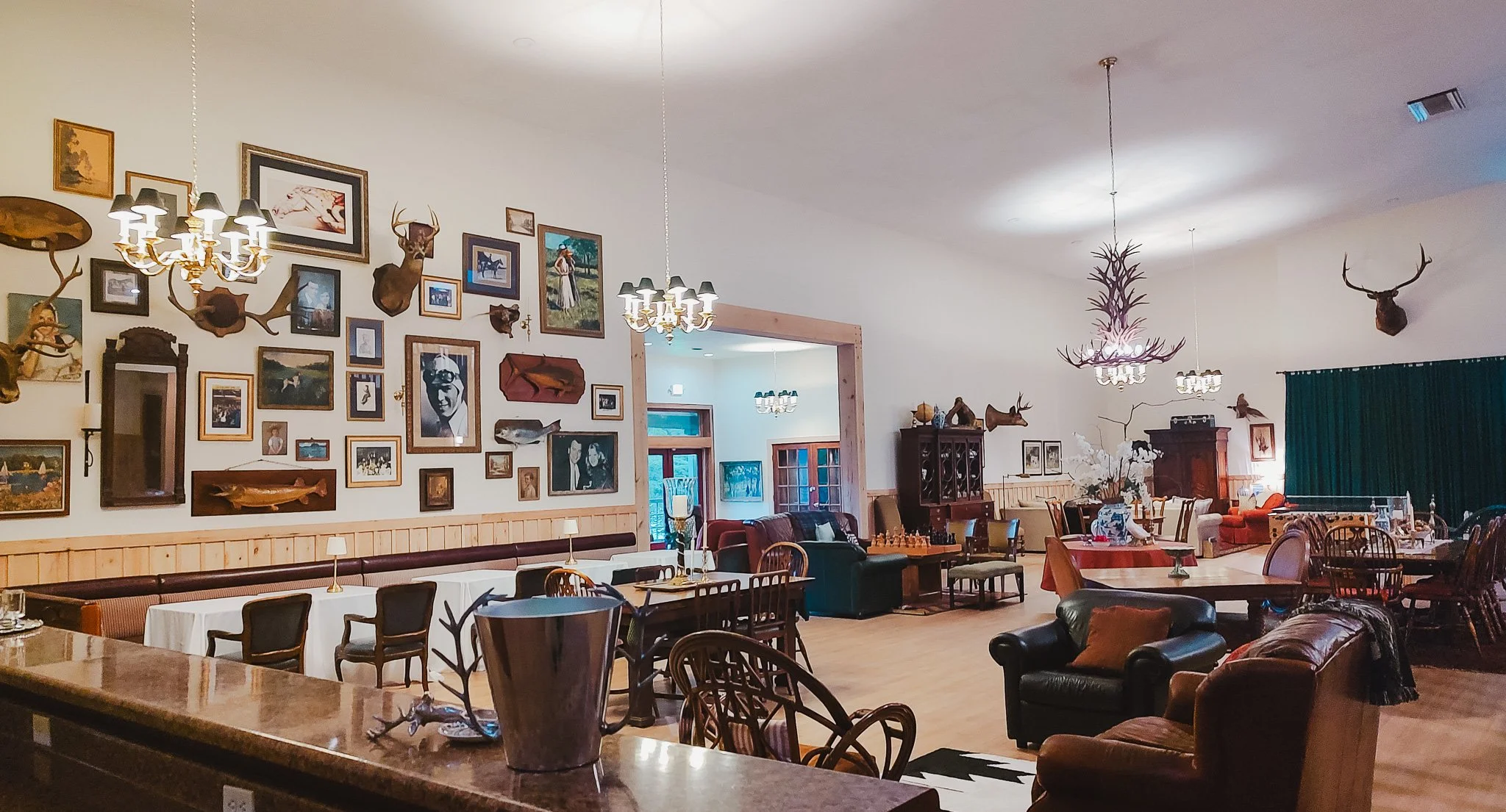 Interior of a spacious lounge or dining area with a high ceiling and woodland-themed decor. The walls are adorned with numerous framed pictures and mounted animal heads. There are multiple chandeliers and antler light fixtures hanging from the ceilin