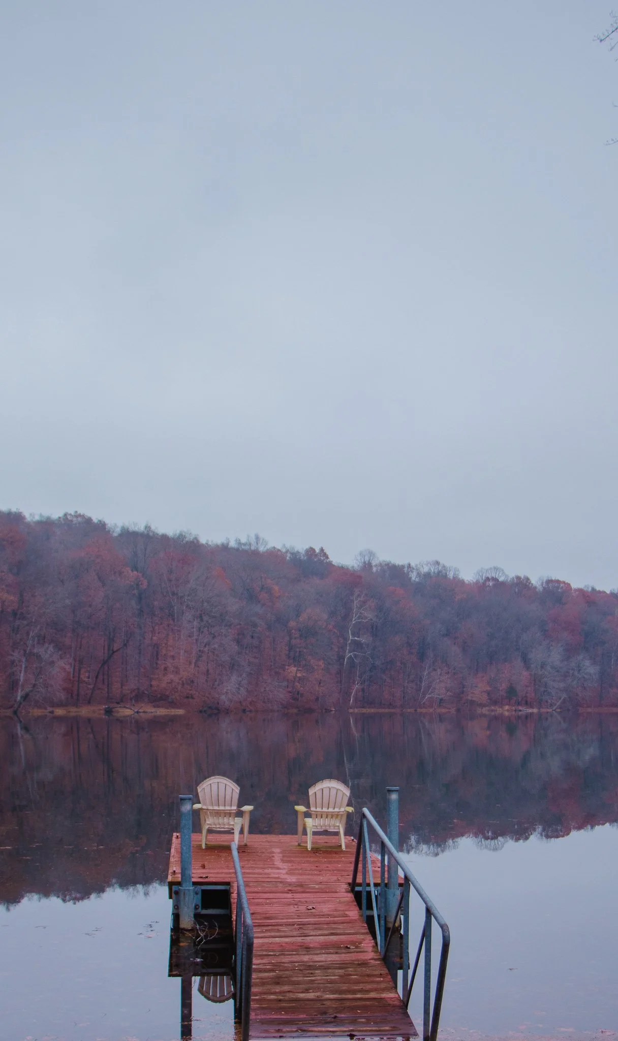 A small wooden dock with two chairs faces a calm lake surrounded by trees with fall foliage, under a cloudy sky.