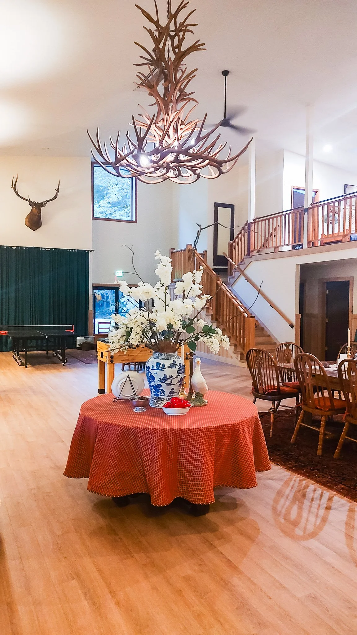 Interior of a spacious room with a round table covered in a red tablecloth, decorated with a large floral arrangement in a blue and white vase, and surrounded by wooden chairs. The room features mounted deer heads on the walls, a staircase with woode