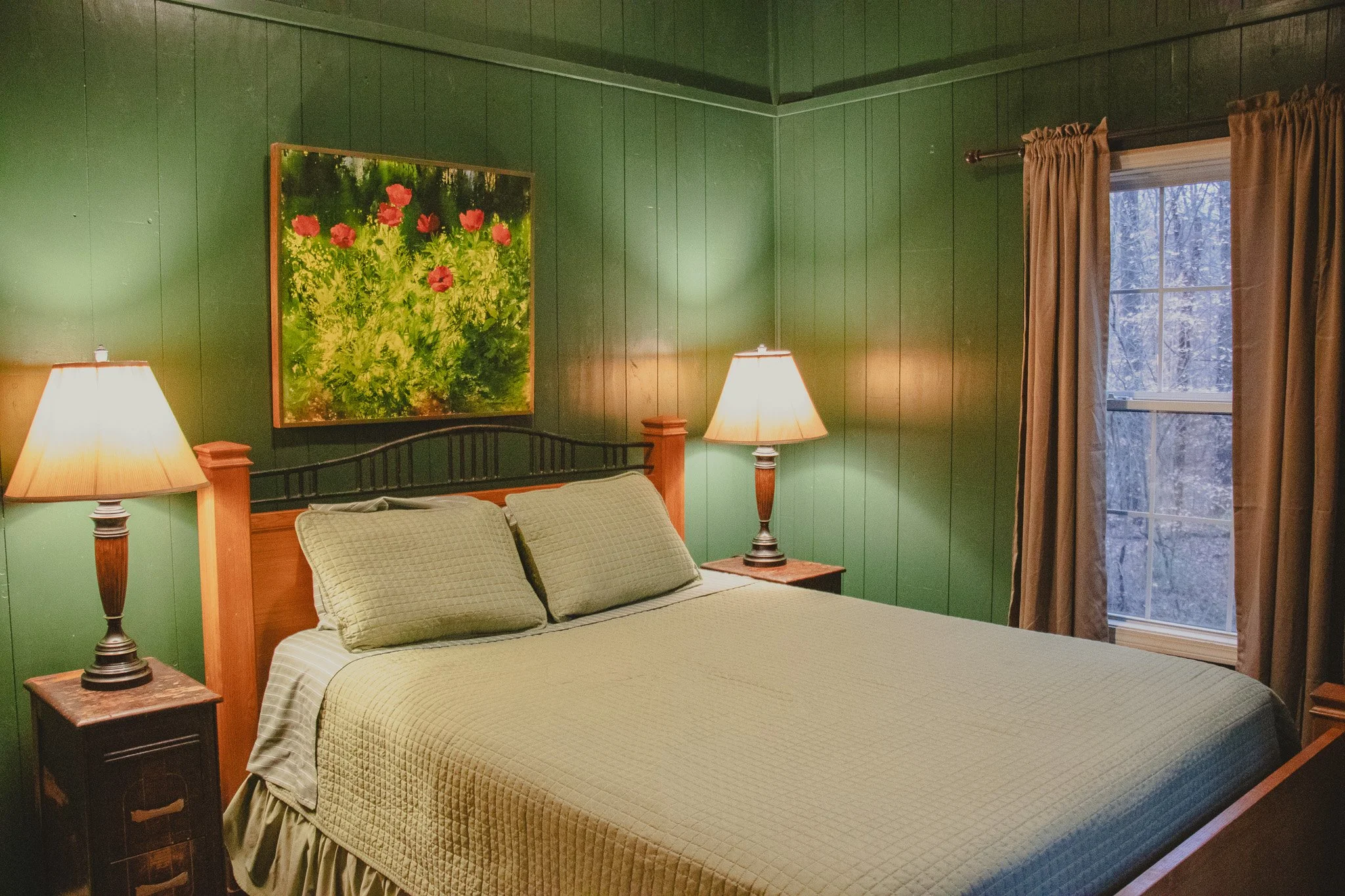 A bedroom with green wooden walls, a bed with beige bedding and pillows, two nightstands with lamps, a painting of pink flowers on the wall, and a window with curtains showing a winter scene outside.