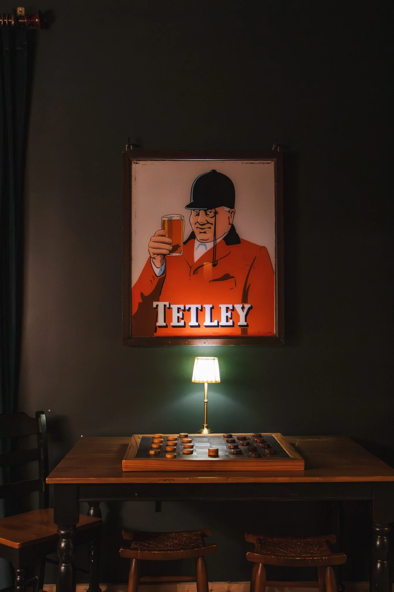 Artistic illustration of a man in a black helmet, orange jacket, and white shirt, holding a glass of beer, with the word 'TETLEY' below. The scene is lit by a small lamp on a wooden table with a vintage board game.