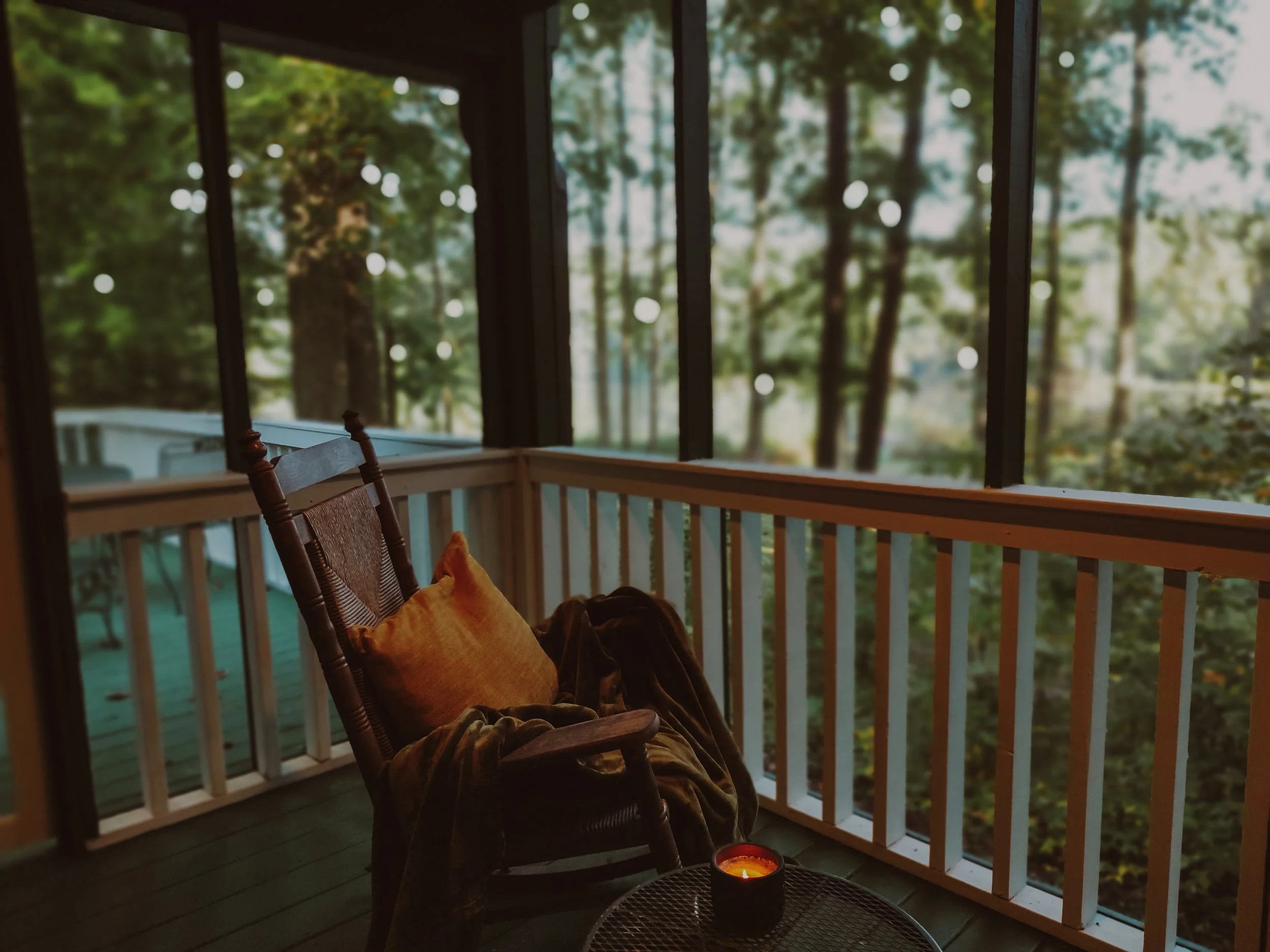 A cozy screened-in porch with a rocking chair draped with a blanket, an orange pillow, a lit candle on a small table, and a view of a wooded backyard with trees and string lights.