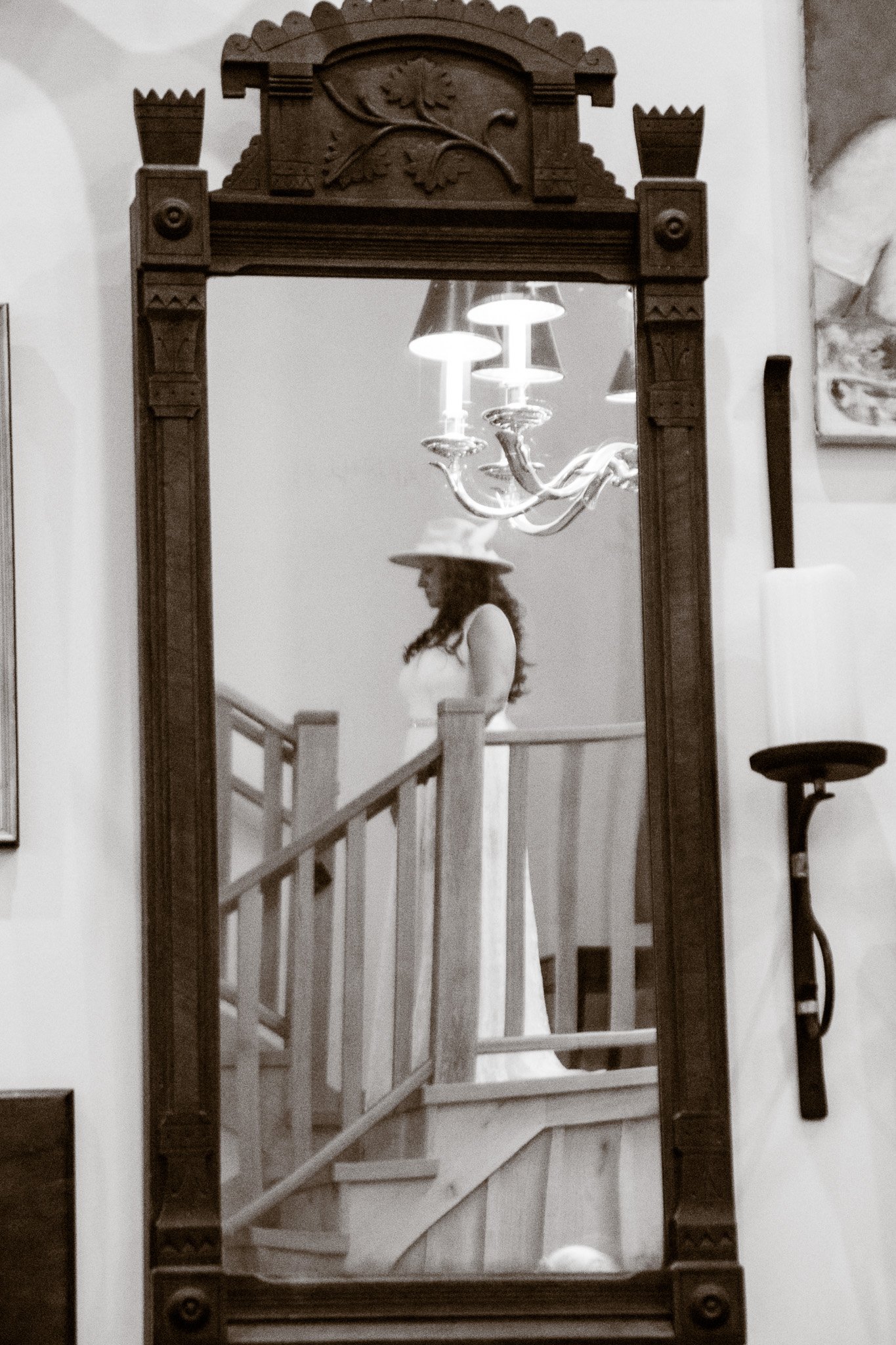 A reflection of a woman in a tall, ornate mirror with a wooden frame featuring carved designs at the top. The woman is wearing a wide-brimmed hat and a long dress, standing on a wooden staircase inside a house with a chandelier hanging from the ceili
