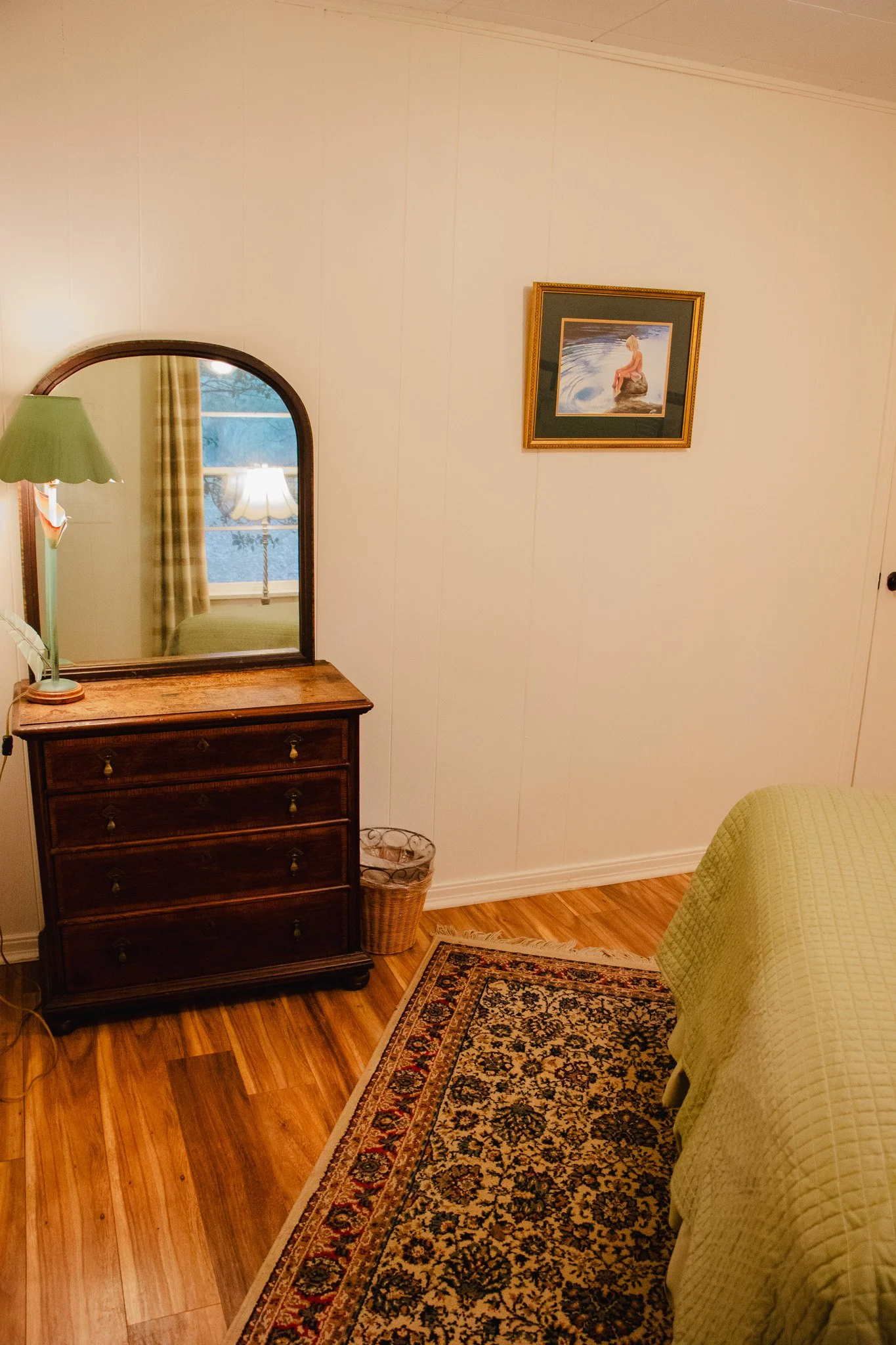 Corner of a bedroom with a wooden dresser with a mirror, a table lamp, a wall art of a woman sitting on a rock by the water, a window with curtains, part of a bed with a yellow cover, and a traditional patterned area rug.