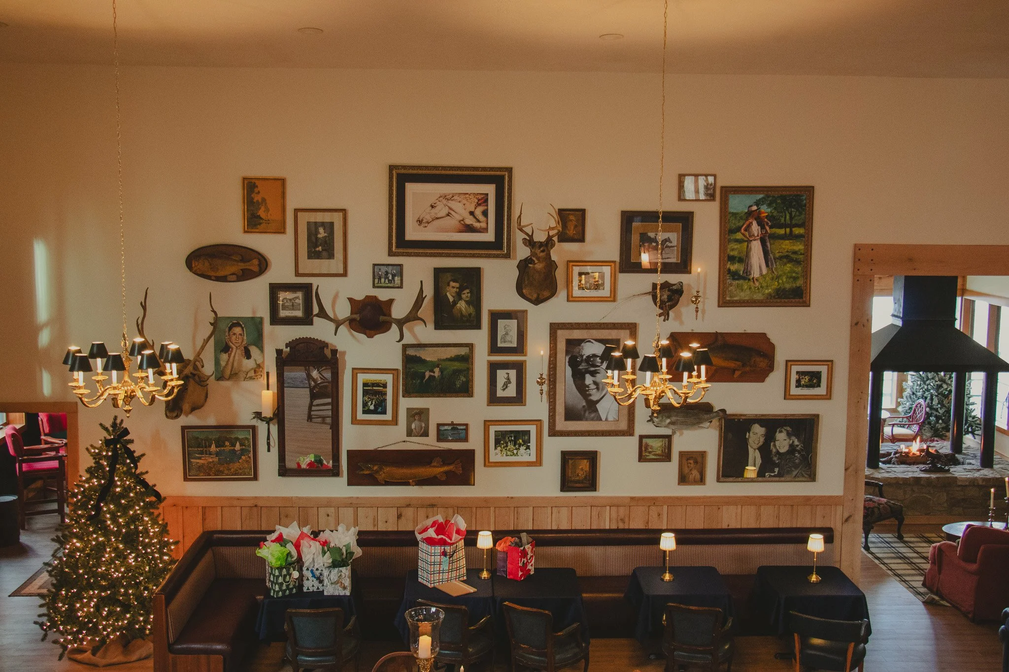 Interior of a cozy, rustic restaurant with a decorated Christmas tree in the corner, and a wall adorned with eclectic framed photos and paintings including animal mounts and a portrait of a man with glasses. There are chandeliers and small table lamp