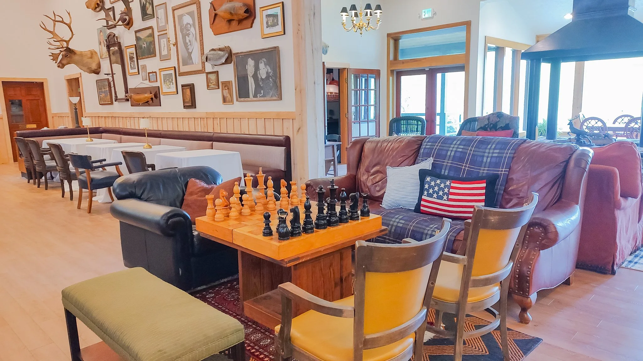 Interior of a cozy lounge or dining area with a large leather sofa, a wooden chessboard, and various chairs. The walls are decorated with framed photos, animal mounts, and artwork. There are several tables with white tablecloths and candles, and a la