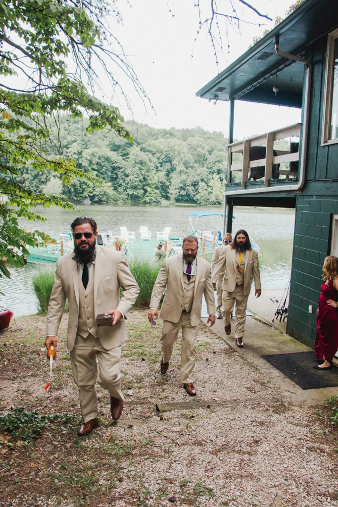 A group of men in beige suits walking along a lakeside path during a wedding, with a green house to the right and a lake and forest in the background.
