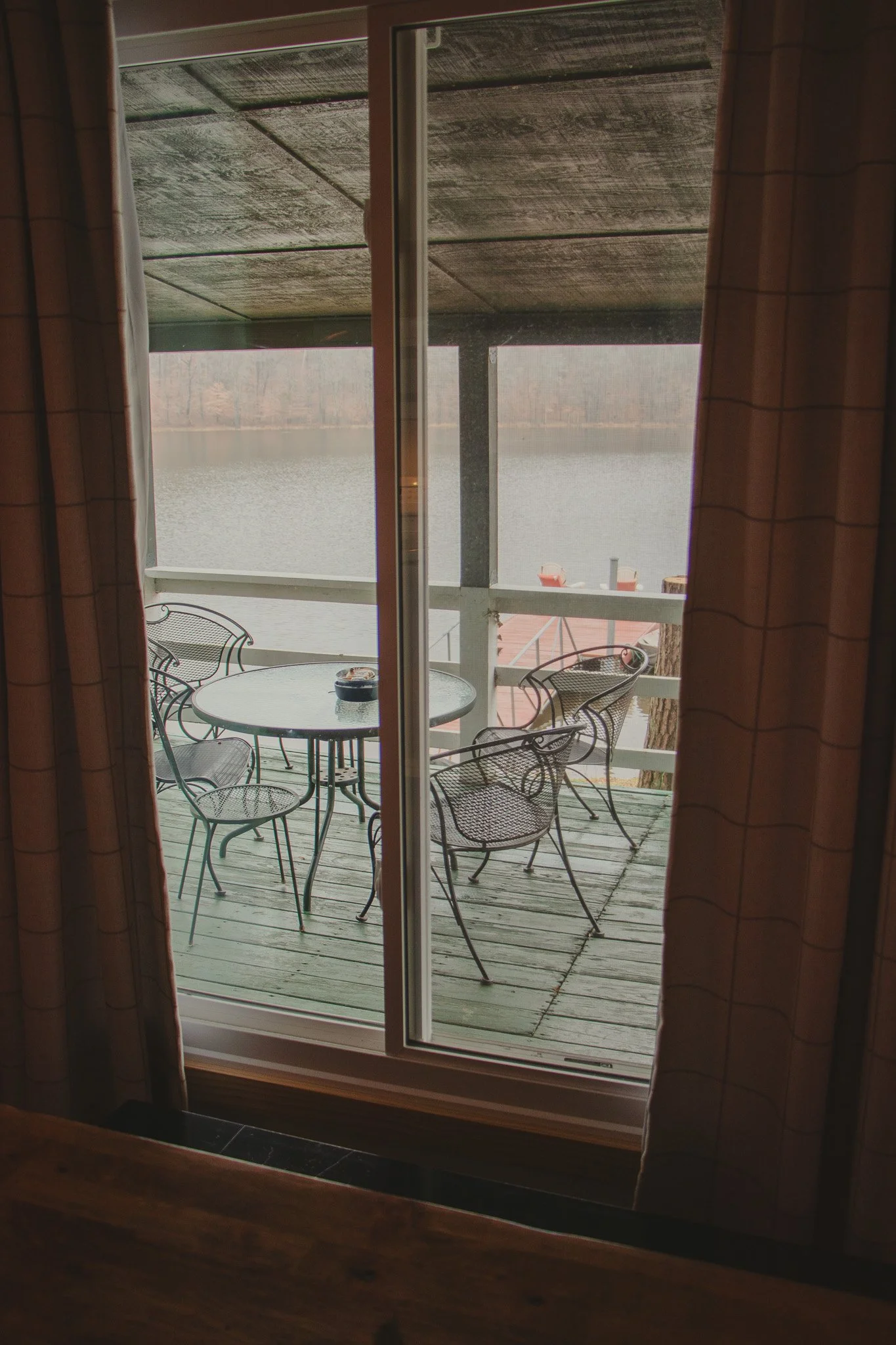View of a lakeside outdoor patio with metal chairs and a table, seen through a sliding glass door from inside a cabin or house.