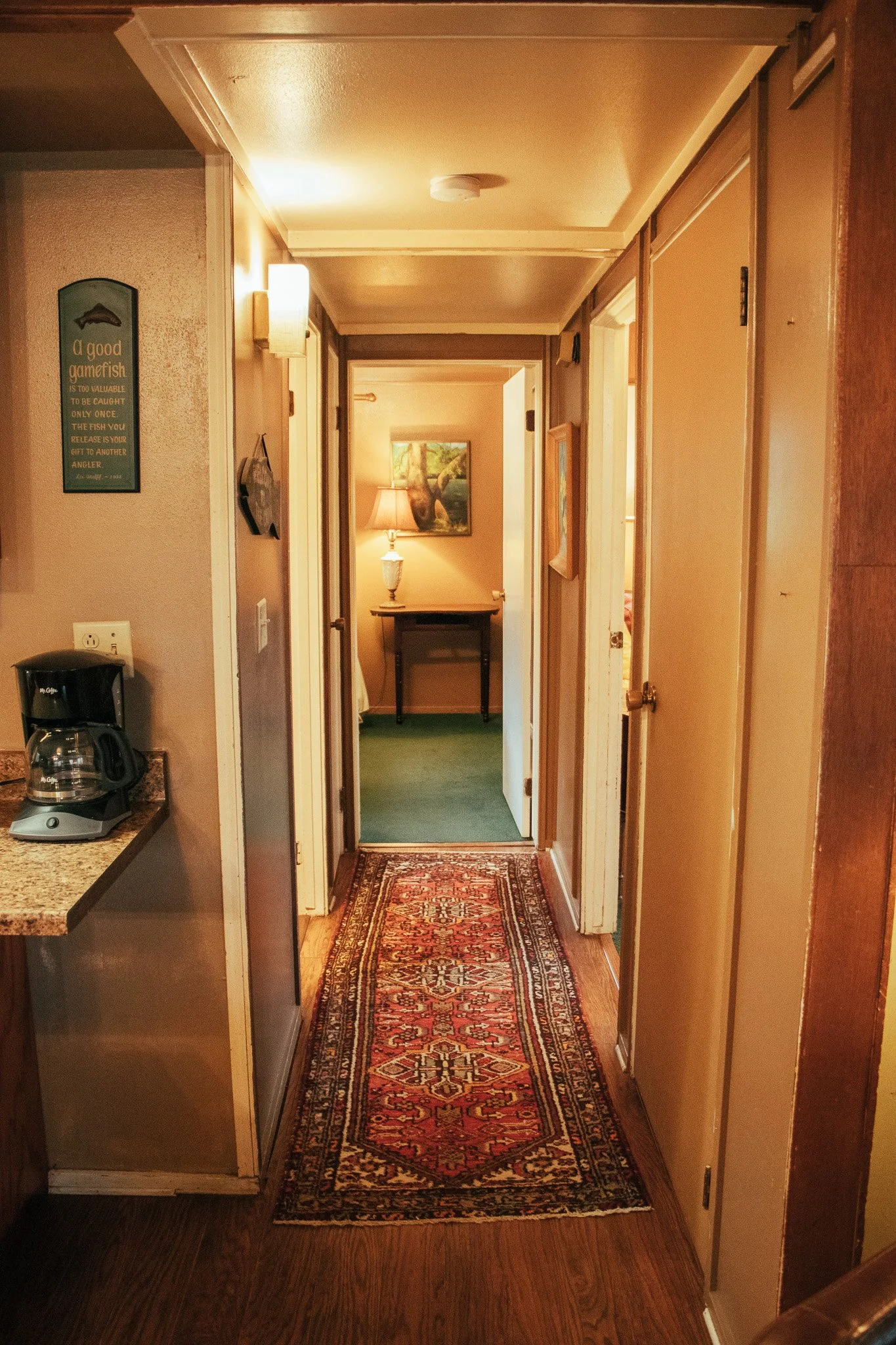 A hallway in a home or hotel with a colorful patterned runner rug, beige walls, and a view into a room with a table, lamp, and artwork.