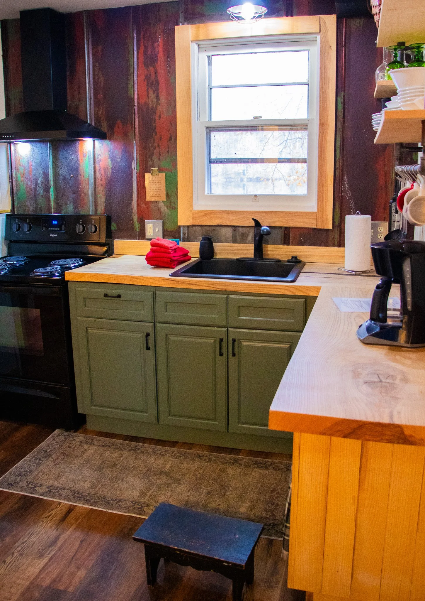A cozy kitchen with a black stove, green cabinets, and a wooden countertop. There is a small step stool, a window above the sink, and various kitchen items like a paper towel roll and coffee maker.