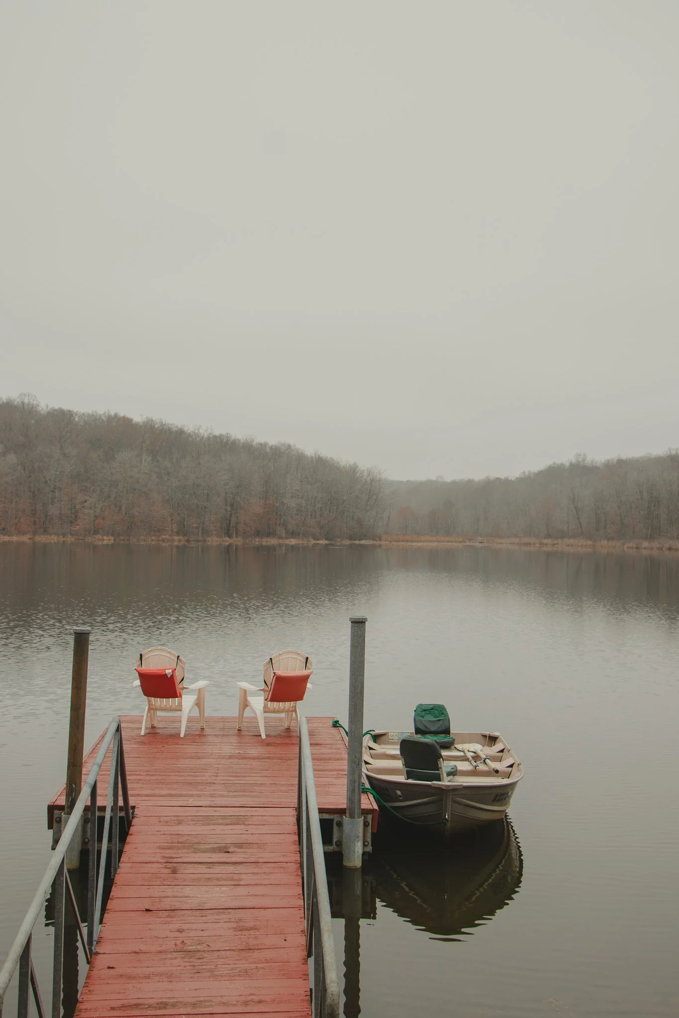 A wooden dock on a calm lake with two orange chairs, a small boat, and a gray overcast sky.