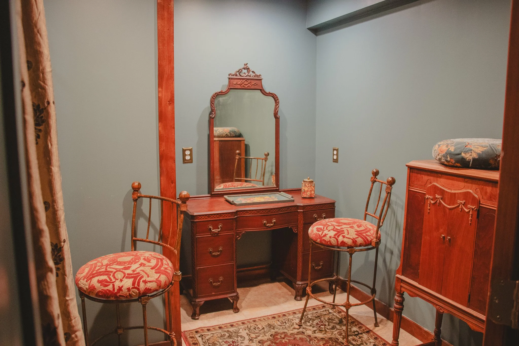 A vintage wooden vanity with a mirror, two matching upholstered chairs, a wooden cabinet with a floral cushion on top, and a patterned rug, all in a room with pale green walls.