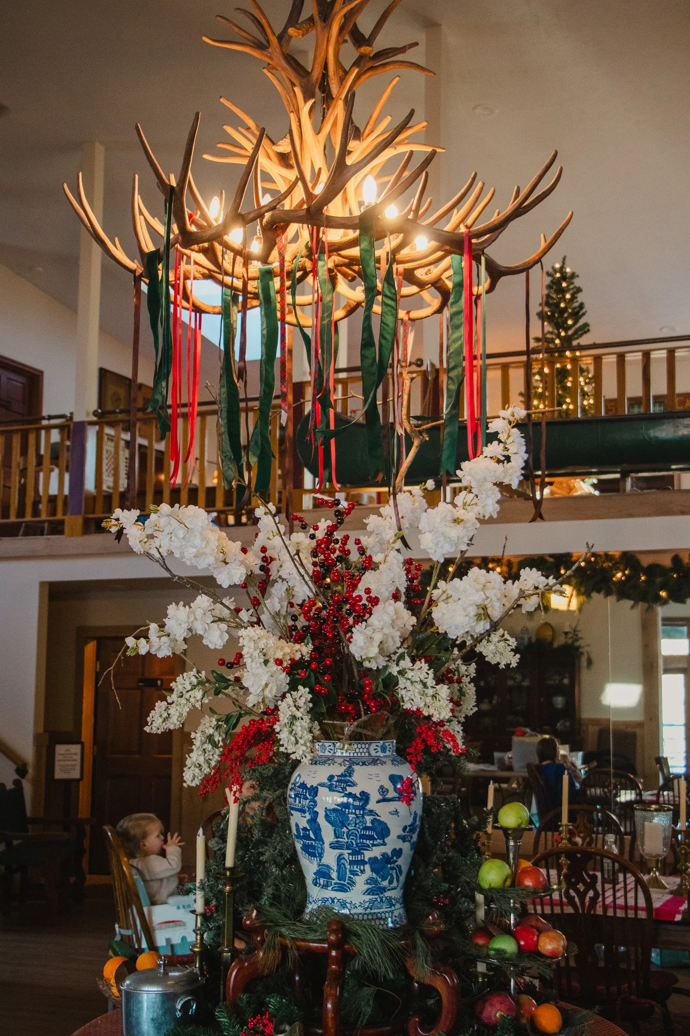 A large floral arrangement in a blue and white vase topped with white flowers and red berries, with a wooden antler chandelier hanging above decorated with ribbons, in a festive indoor setting.