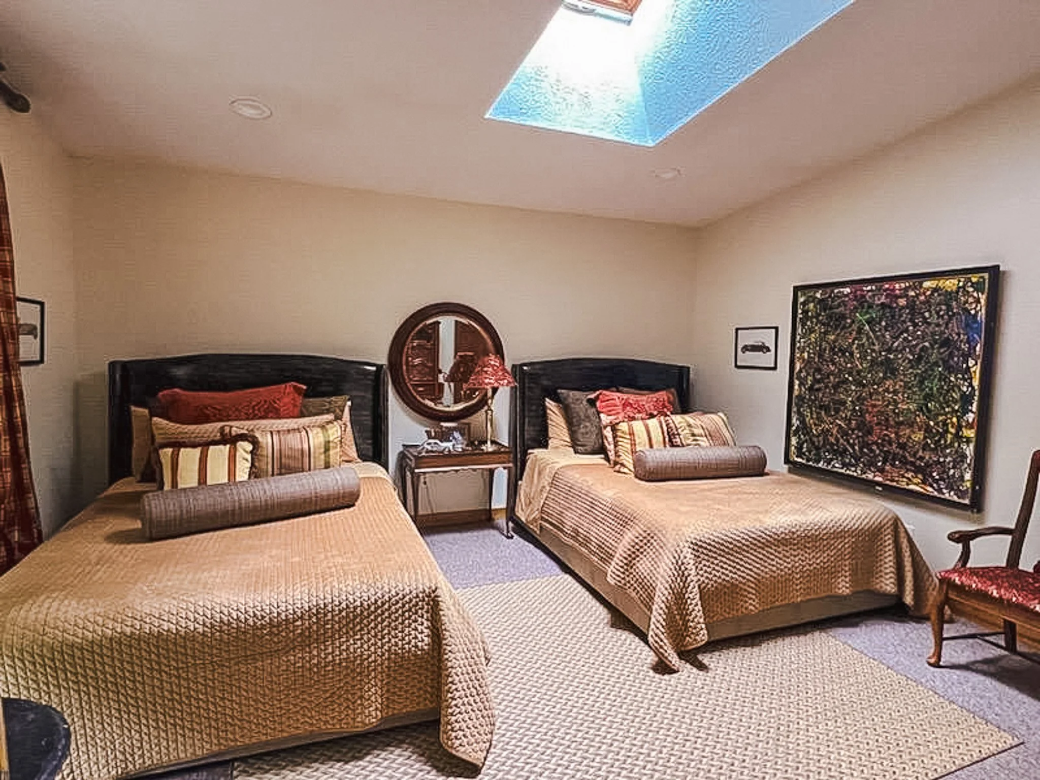 A bedroom with two twin beds, each with a black headboard and tan quilted bedspreads. Between the beds is a wooden nightstand with a lamp that has a patterned shade. Above the nightstand is a round mirror on the wall. The room has a skylight, allowin