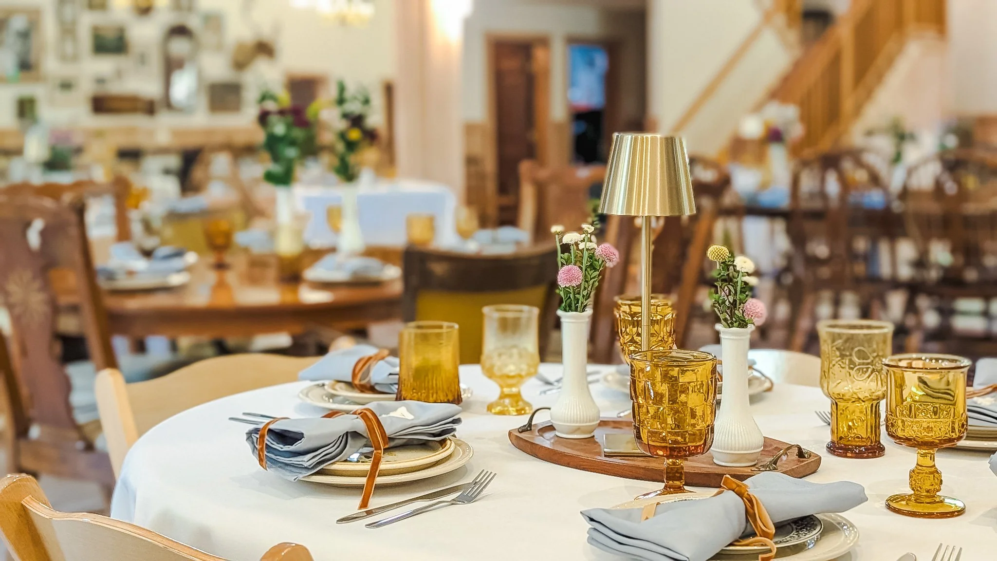 A dining table set with plates, glasses, napkins, and vases with pink and white flowers, in a warmly lit restaurant with wooden furniture.