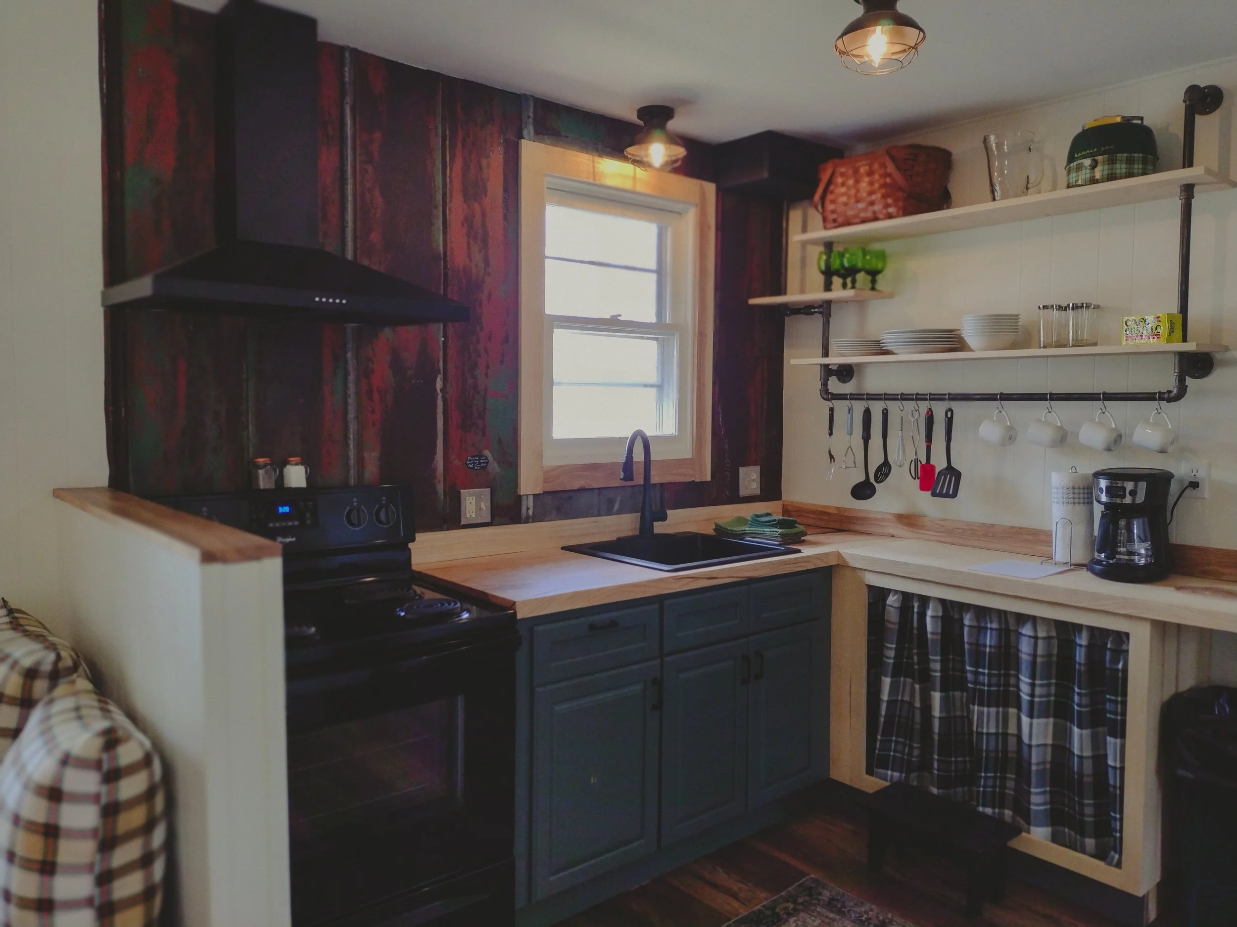 Cozy kitchen with a rustic theme, featuring a black stove, a wooden countertop, dark green cabinets, open shelving with plates, bowls, glasses, and cups, a window above the sink, hanging utensils, a coffee maker, and a plaid curtain under the sink.