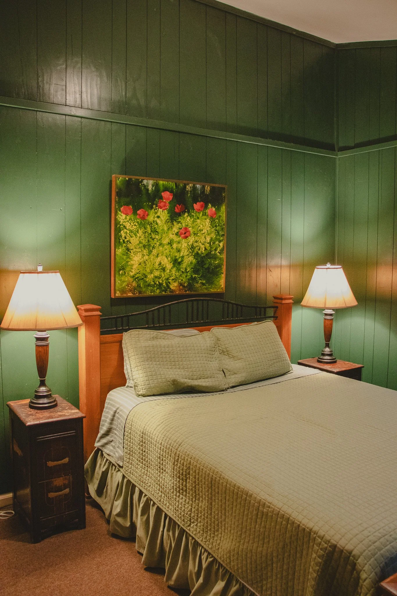 A bedroom with green paneled walls, a bed with a green quilt and matching pillows, wooden bedside tables with lamps, and a framed floral painting on the wall.