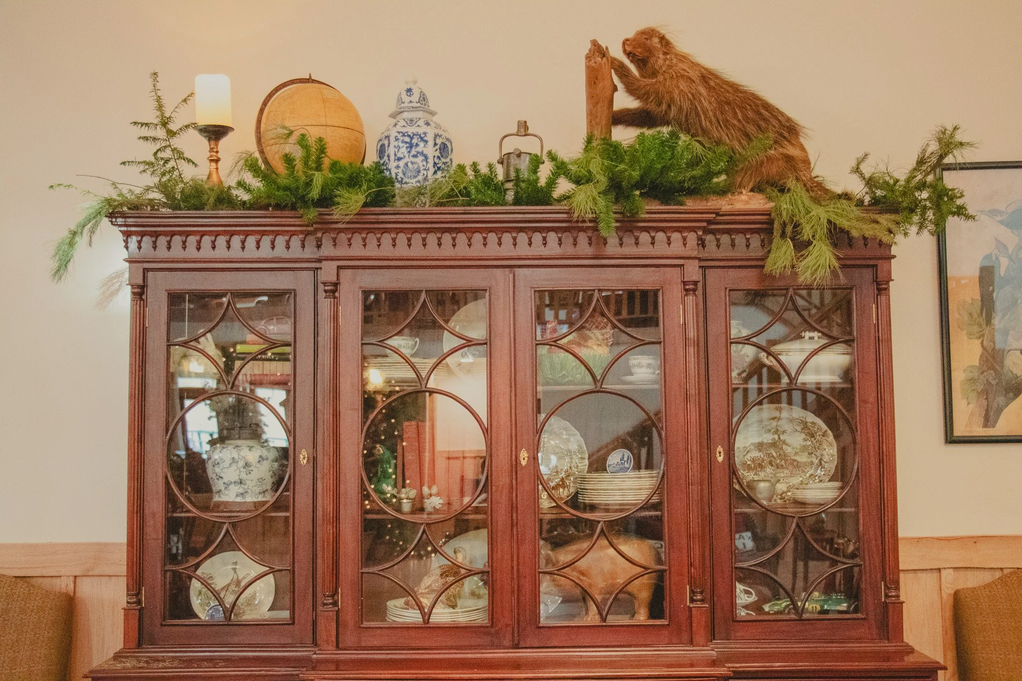 A wooden china cabinet with glass doors displaying plates and dishware inside, decorated with greenery on top, along with a candle, globe, jar, and a fur pelt on top