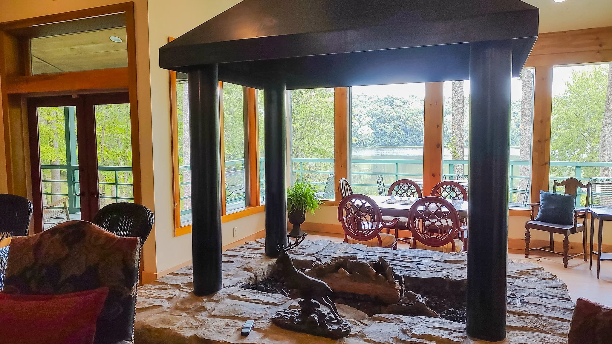 Interior of a house with a stone fireplace, wooden beams, and large windows showing an outdoor balcony with trees and water in the background. There are chairs, a table, and decorative pillows.