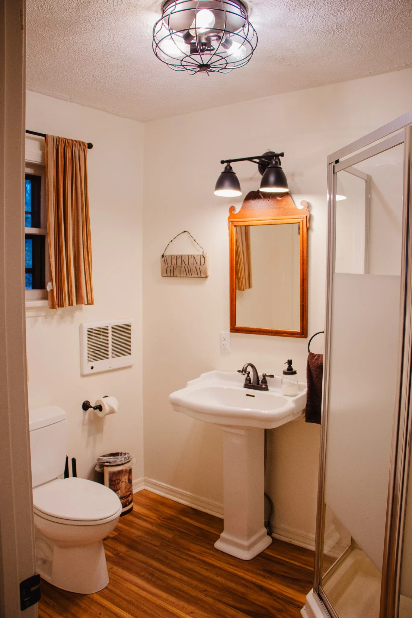 A small bathroom with a white toilet, a pedestal sink with a mirror above, a small window with striped curtains, a wall sign that says 'Weekend Getaway,' a night light fixture, a shower with a sliding door, and wood-look flooring.
