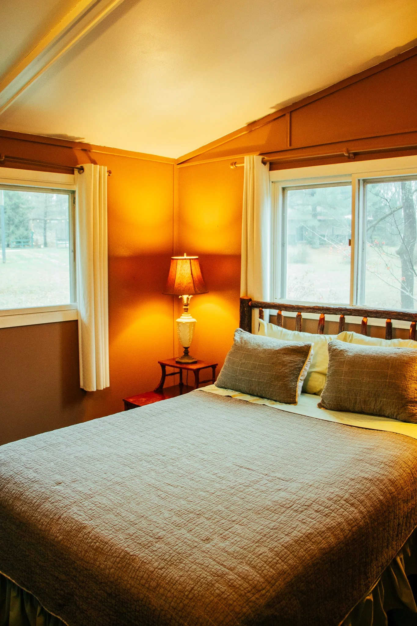 A cozy bedroom with a bed, two pillows, a wooden headboard, a bedside table, a lamp, white curtains, and windows showing an outdoor scene.