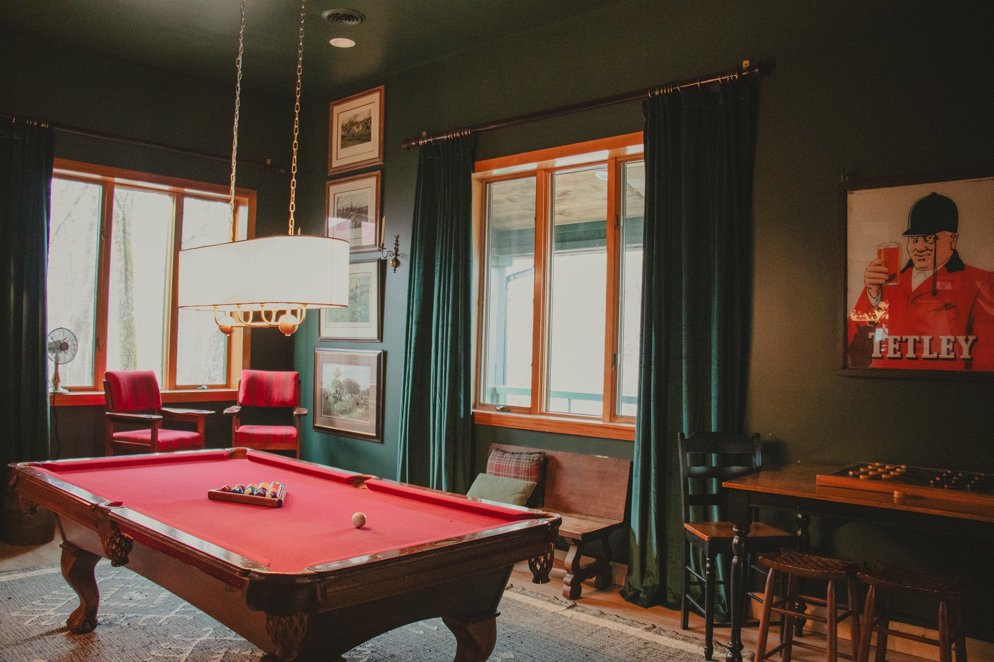 A cozy room with a red pool table, red chairs by the window, green curtains, framed artwork on green walls, and a vintage Tetley poster on the wall.