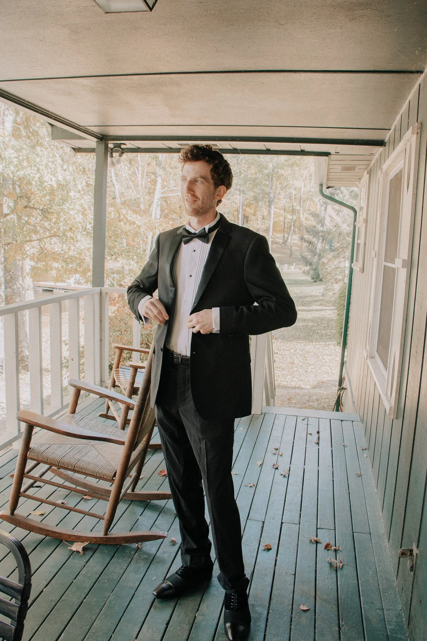A man in a tuxedo adjusting his jacket on a porch with wooden chairs and autumn leaves.