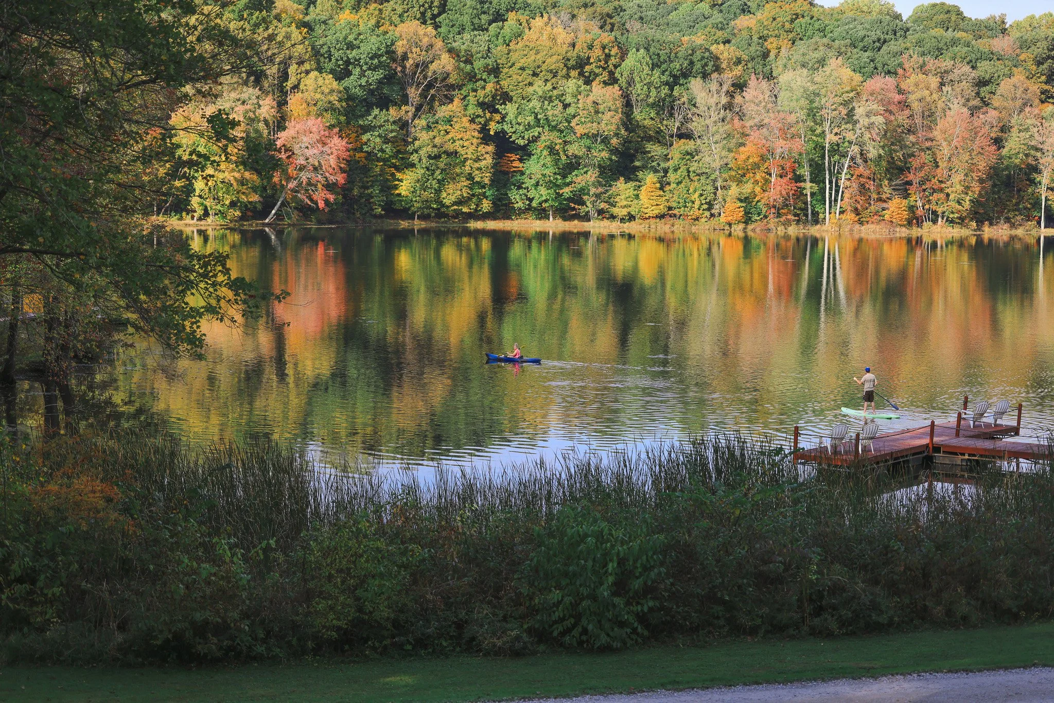 A lake surrounded by trees with fall foliage, two people paddleboarding on the water, and a small dock with chairs on the right side.