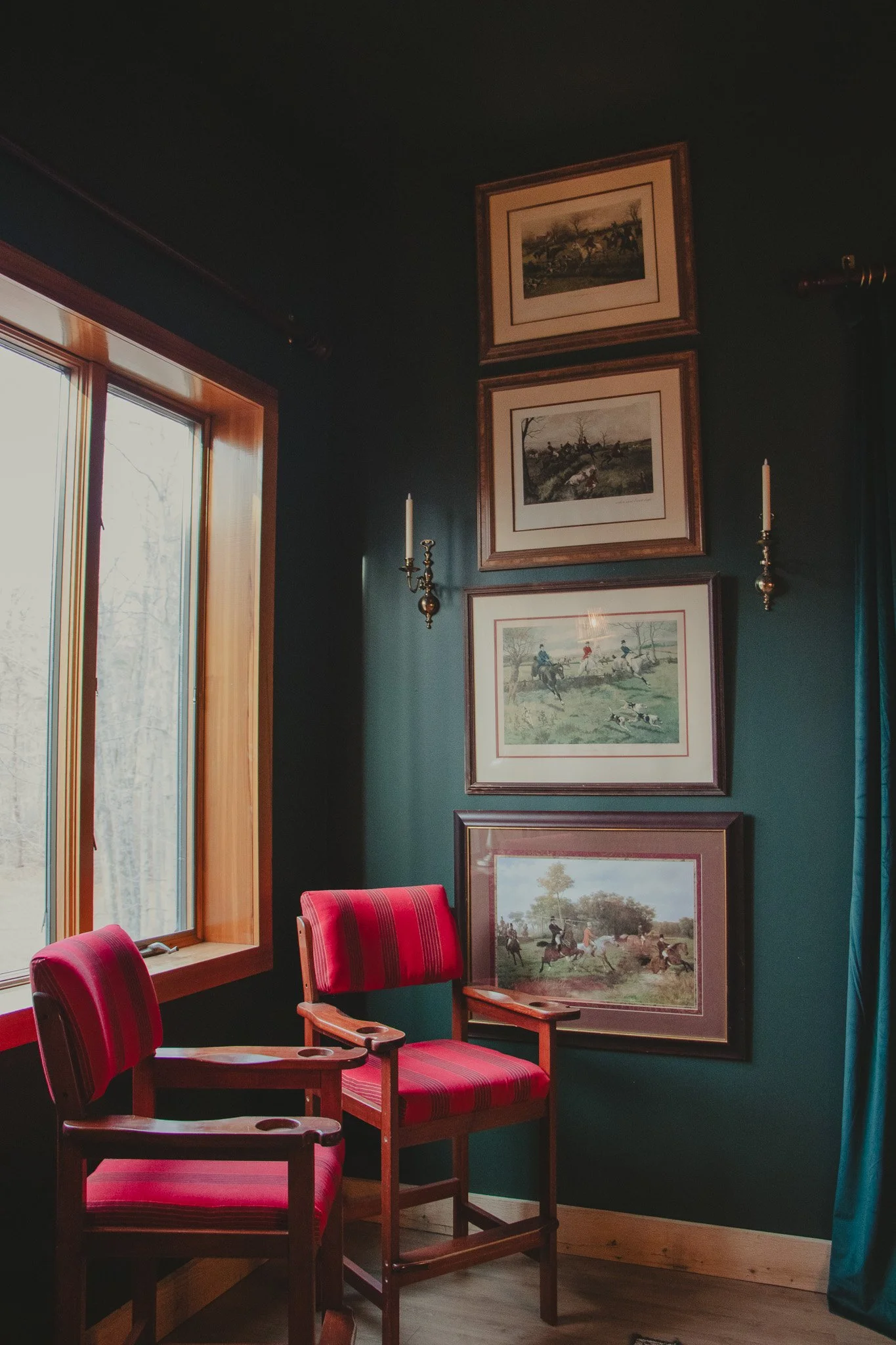 A corner of a room with two red-striped cushioned chairs in front of a large window with wooden trim, dark green walls, and five framed paintings or prints of scenes involving horses and riders, arranged vertically. Two wall-mounted candle sconces ar