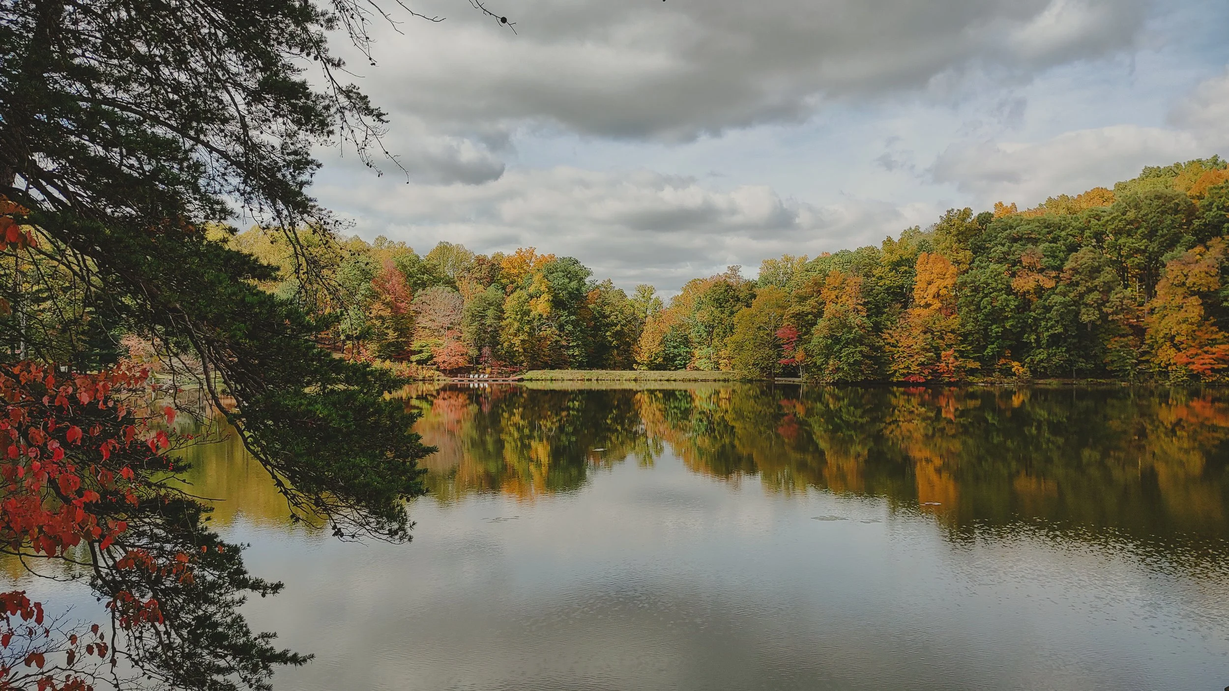 A calm lake reflecting the colorful autumn trees surrounding it, with overcast sky above.