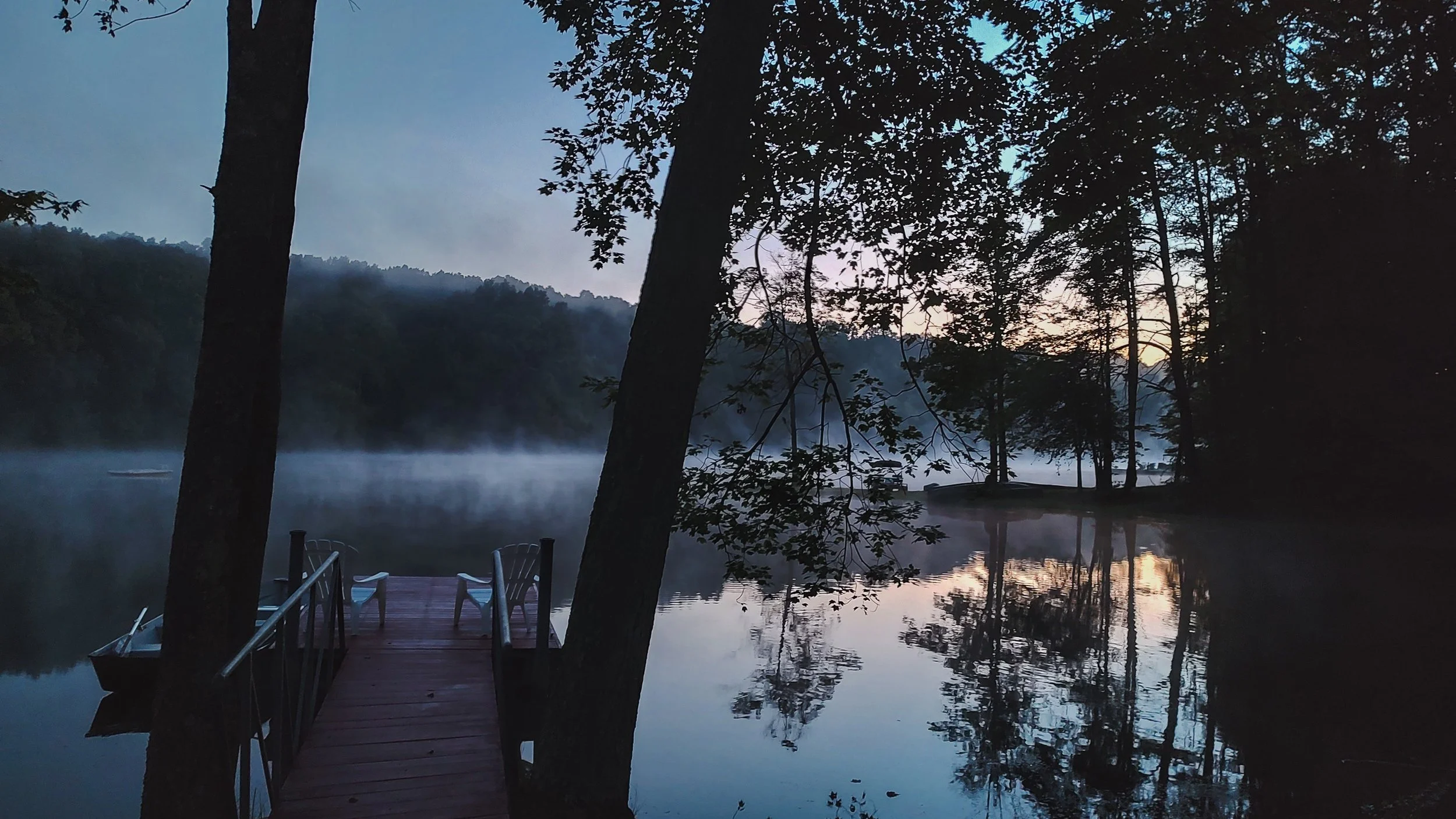 A wooden dock with two chairs is on a calm lake surrounded by trees at dusk. Mist rises from the water, and the sky is starting to darken, reflecting the silhouettes of the trees.