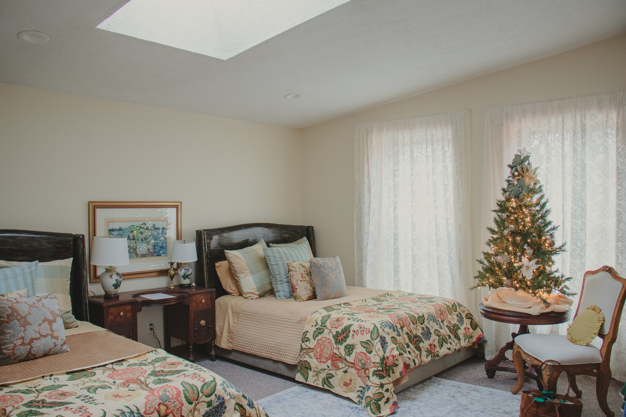 Two beds with floral bedspreads and pillows in a bedroom decorated for Christmas. A small Christmas tree with lights and ornaments stands next to a white chair with a yellow pillow. There are windows with sheer lace curtains, and a framed picture han