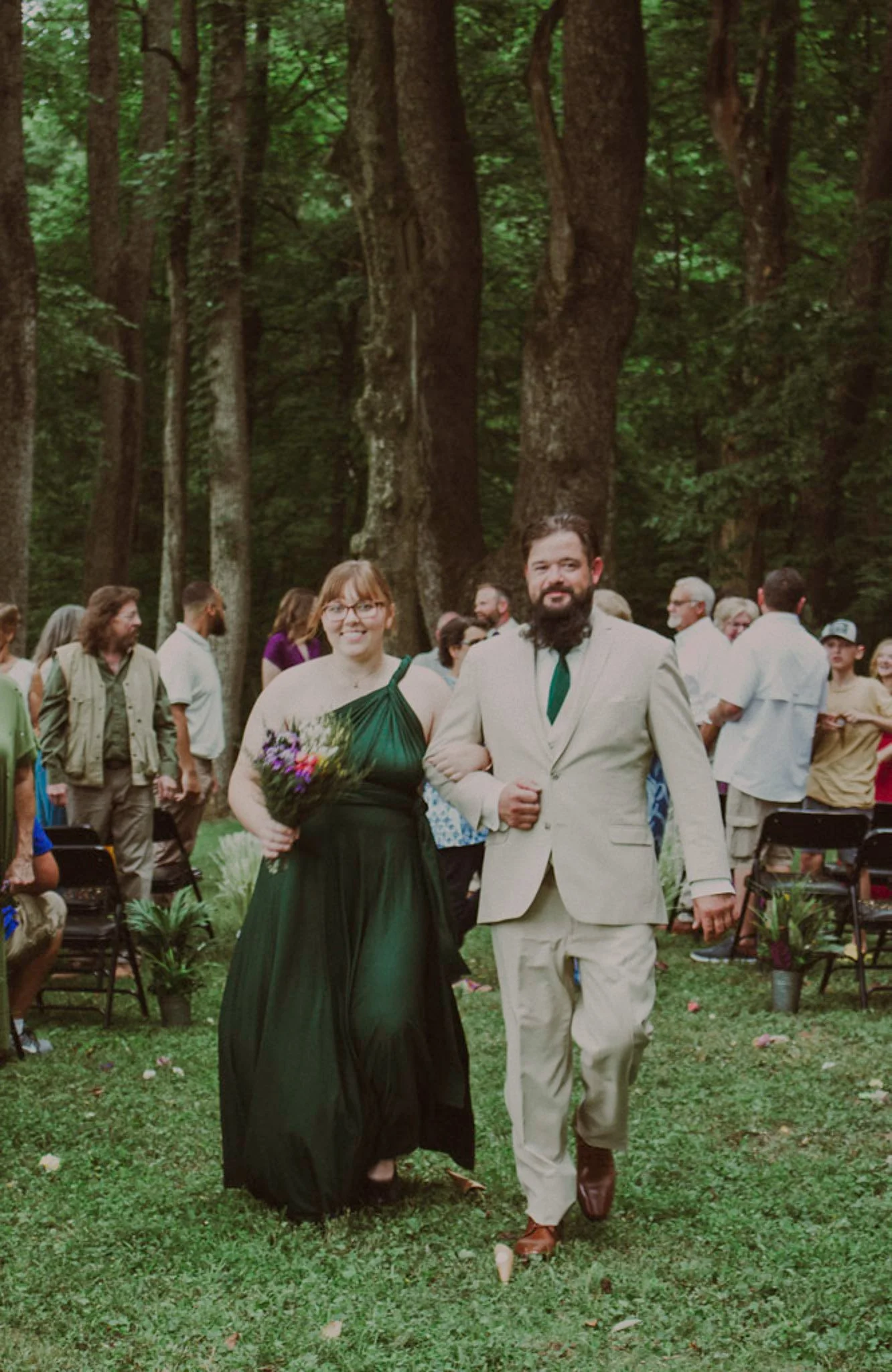 A wedding ceremony outdoors in a wooded area, with a woman in a green dress holding a bouquet and a man in a beige suit walking down the aisle, surrounded by guests.