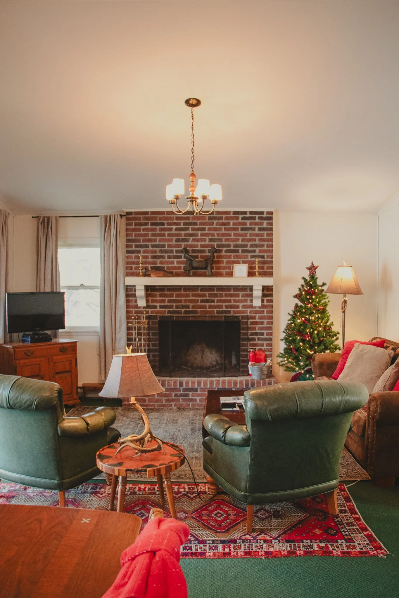 Living room decorated for Christmas with a small Christmas tree adorned with lights and ornaments, a brick fireplace with mantle, a sofa with cushions, two green armchairs, a side table with a deer antler lamp, a television, and a window with curtain