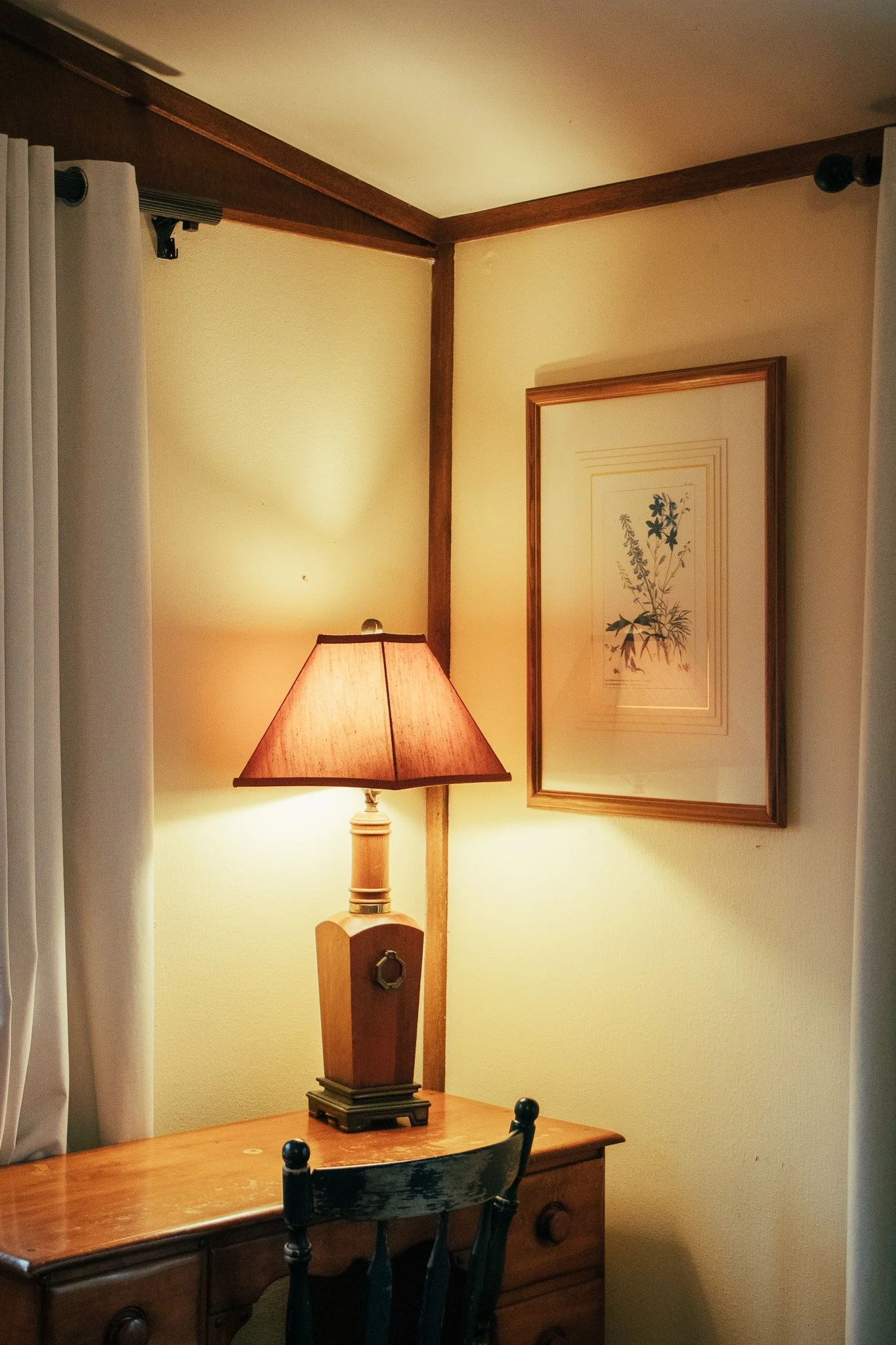 A corner of a room with a wooden desk, a table lamp with a pinkish shade, framed botanical artwork on the wall, a curtain on a window, and a wooden chair.