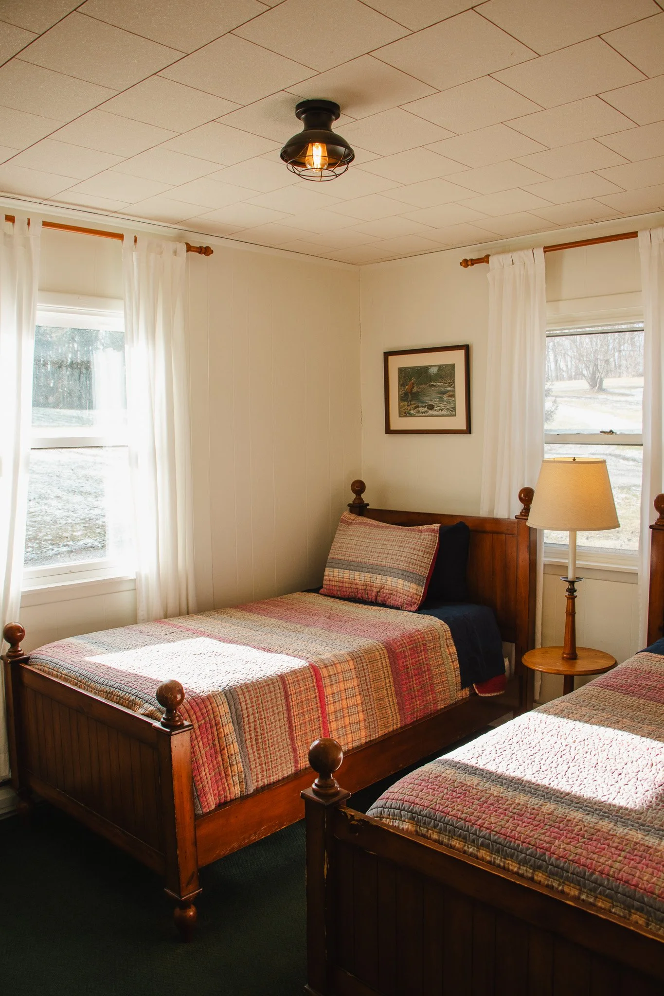 A cozy bedroom with two twin beds with plaid bedding, a wooden nightstand with lamp, and windows with white curtains.