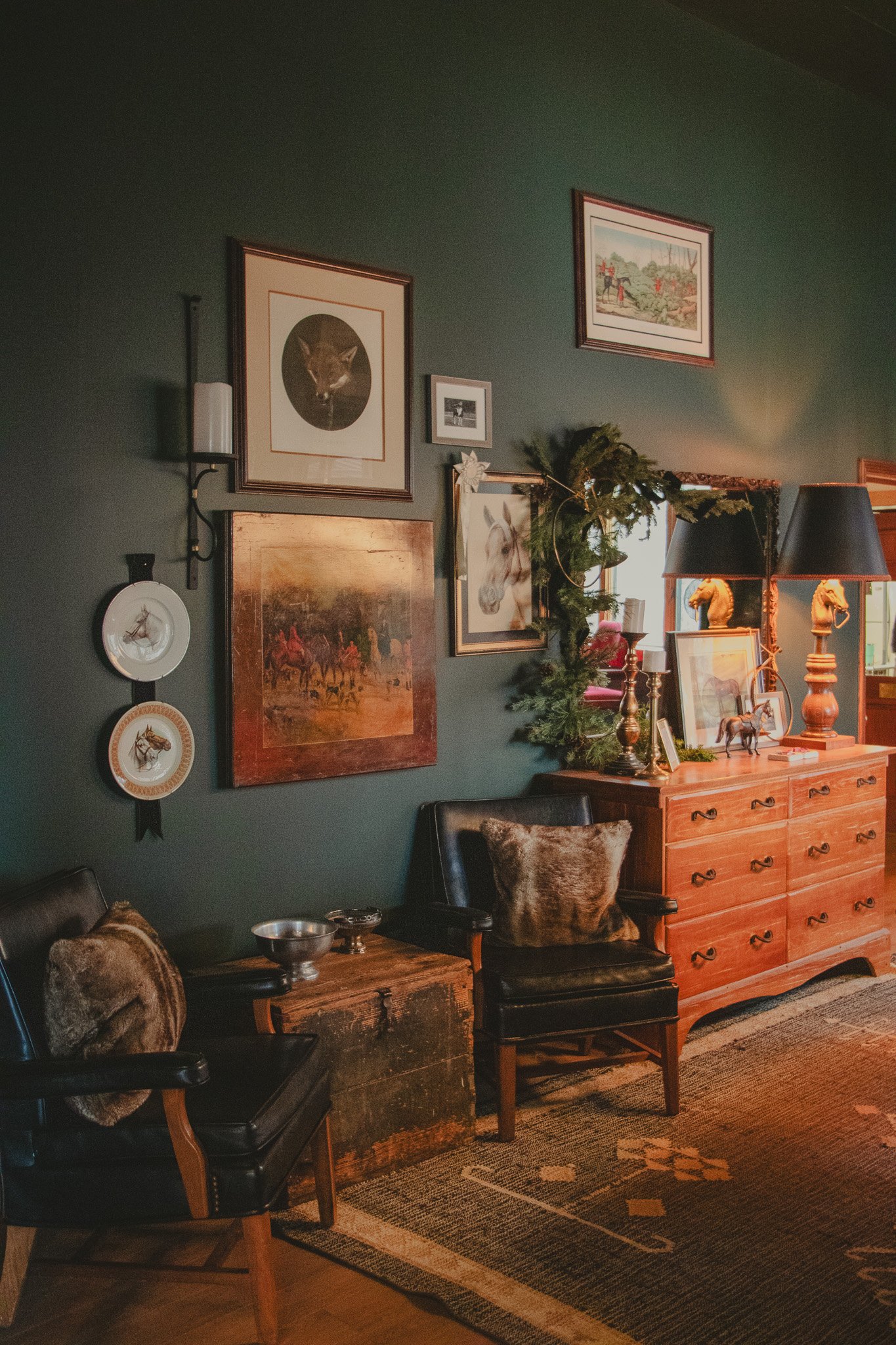Interior of a living room with green walls, vintage furniture, framed artwork including a cat, horse, and polar bear, decorative plates, and a dark wood dresser with lamps and holiday greenery.