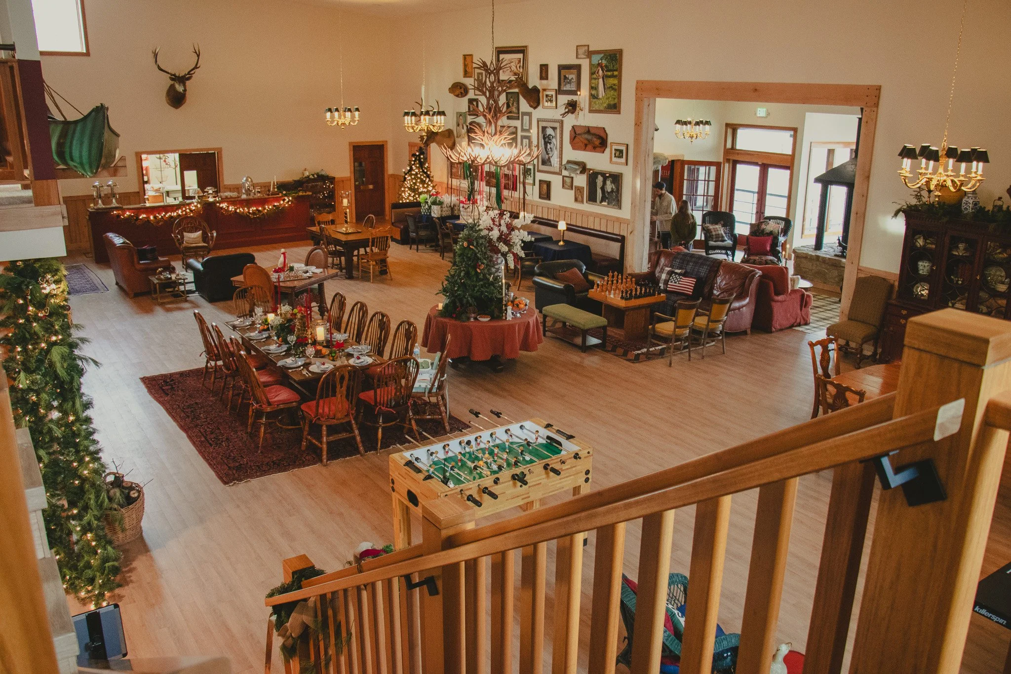 A spacious, decorated living room with festive holiday decorations, a decorated Christmas tree, a large Christmas centerpiece, and various seating areas, including a long dining table set for a meal, a foosball table, and multiple couches and chairs.