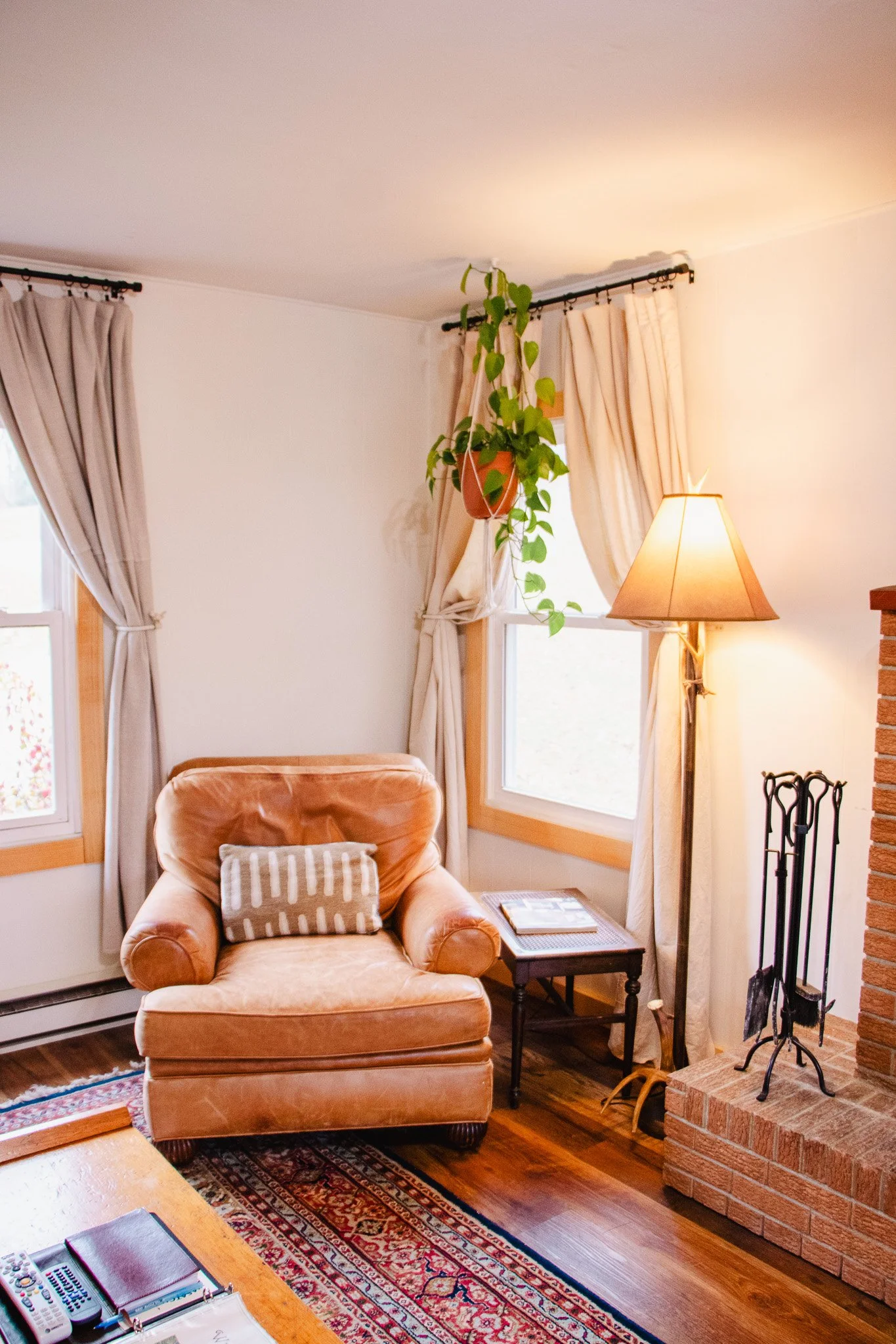 Living room corner with an orange armchair, side table, and a floor lamp. Two windows with beige curtains, a hanging plant, and a brick fireplace with fireplace tools.